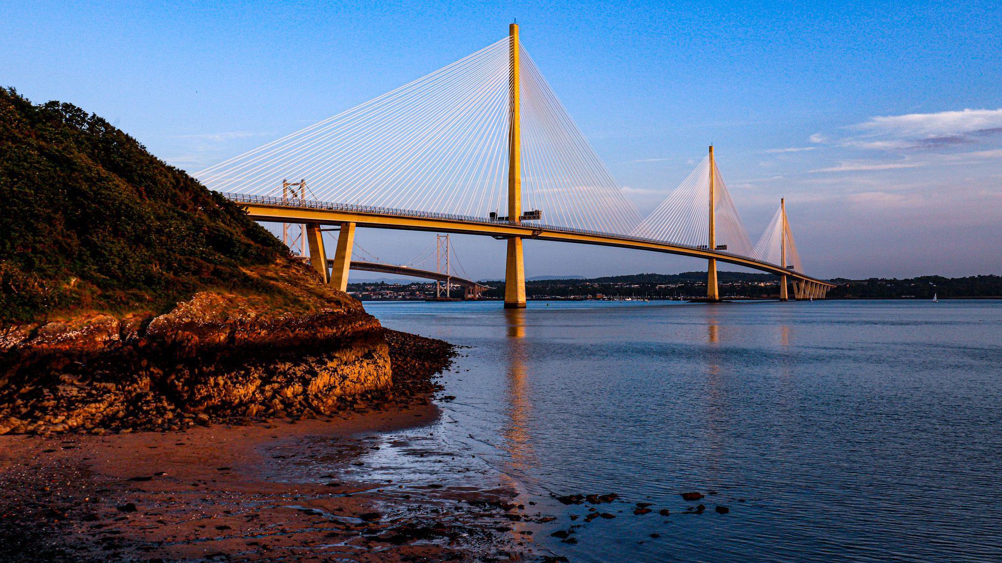 The Queensferry Crossing, which is white, going across the Firth of Forth below. The picture is taken at sunset and there is a gold glow against the bridge. The foreground has a small beach and water in it with a small hill with a patch of green.