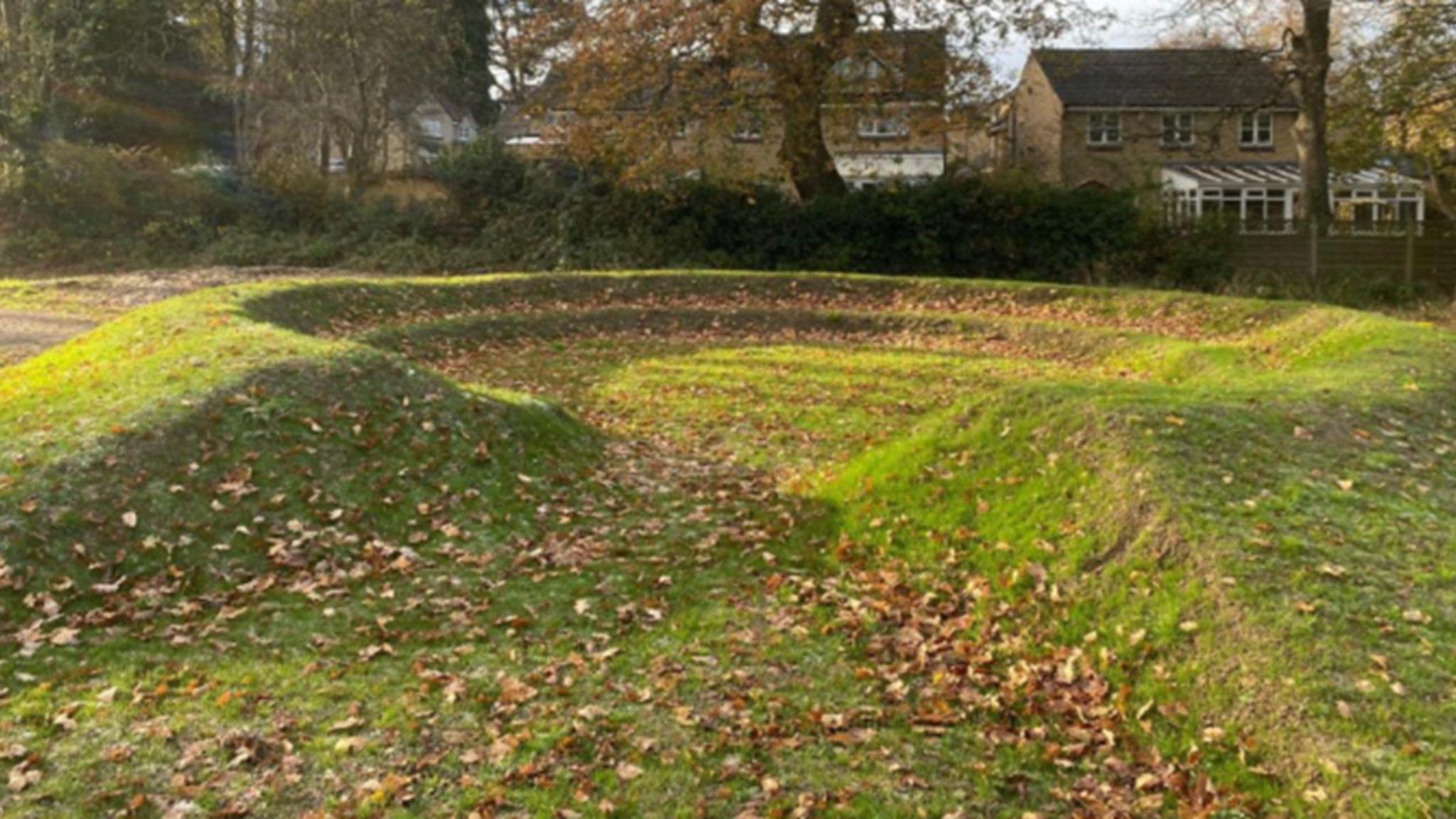 A small patch of grassland which has been manipulated to look like a very small amphitheatre with two rows. Autumn leaves litter the ground. Some houses and trees are in view in the background.