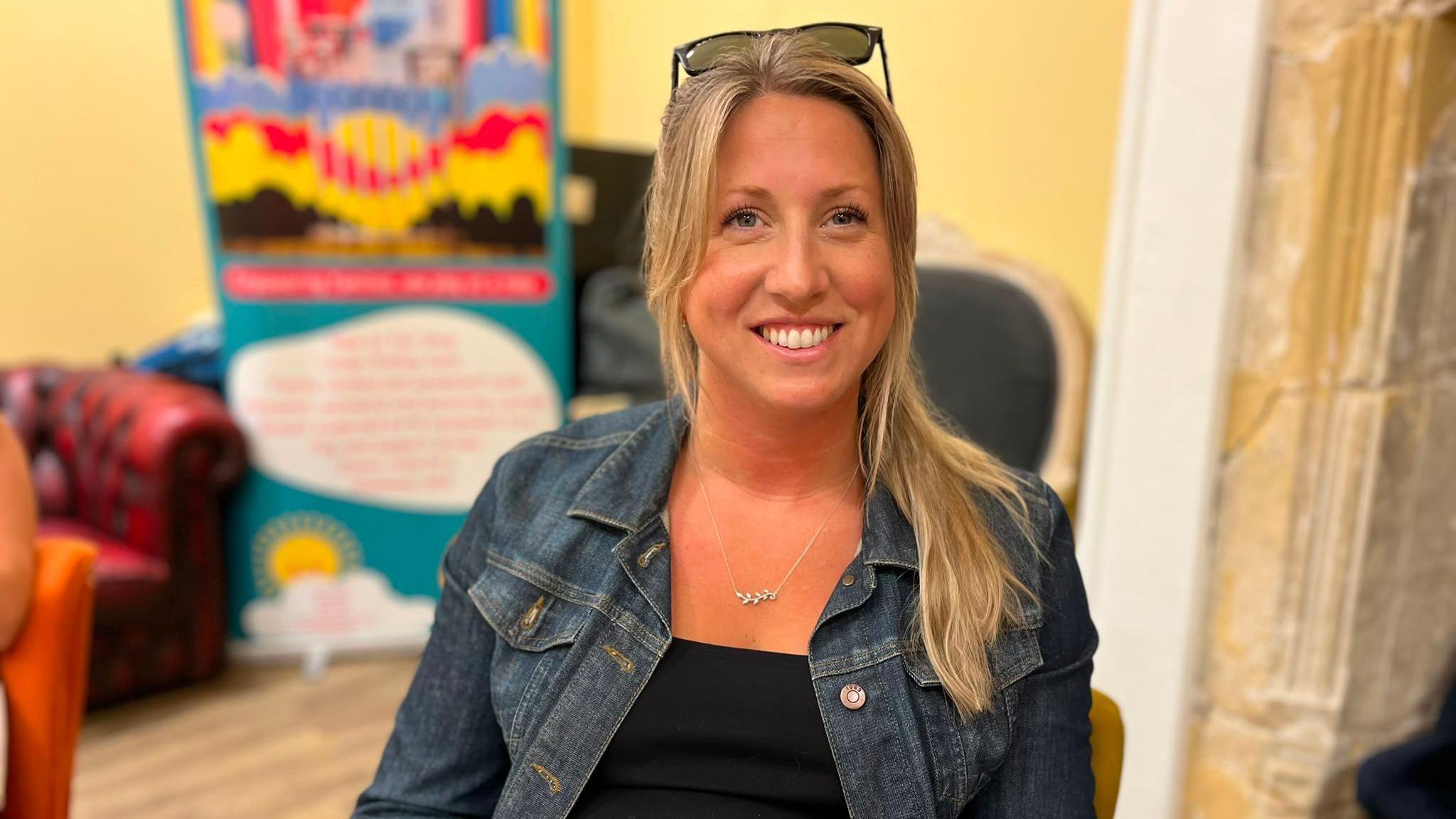 Hannah Winnie-Dennis smiling at the camera. She had blonde hair partly tied up and sunglasses on her head. She's sat in front of a colourful sign.