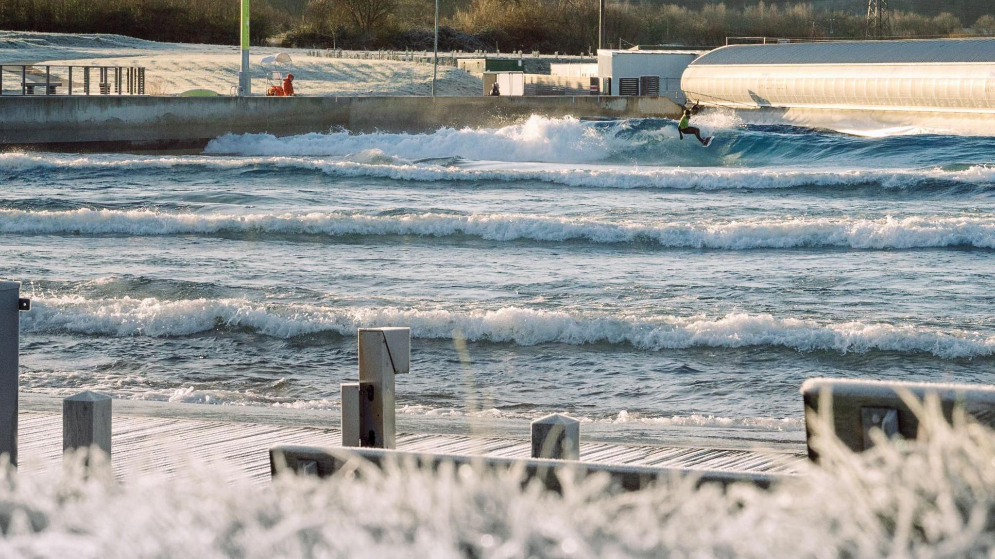 A surfer rides a wave in the distance at the man made surfing lake The Wave on the outskirts of Bristol. In the foreground heavily-frosted grass is visible, while between the surfer and the camera are several waves travelling across the surface of the water