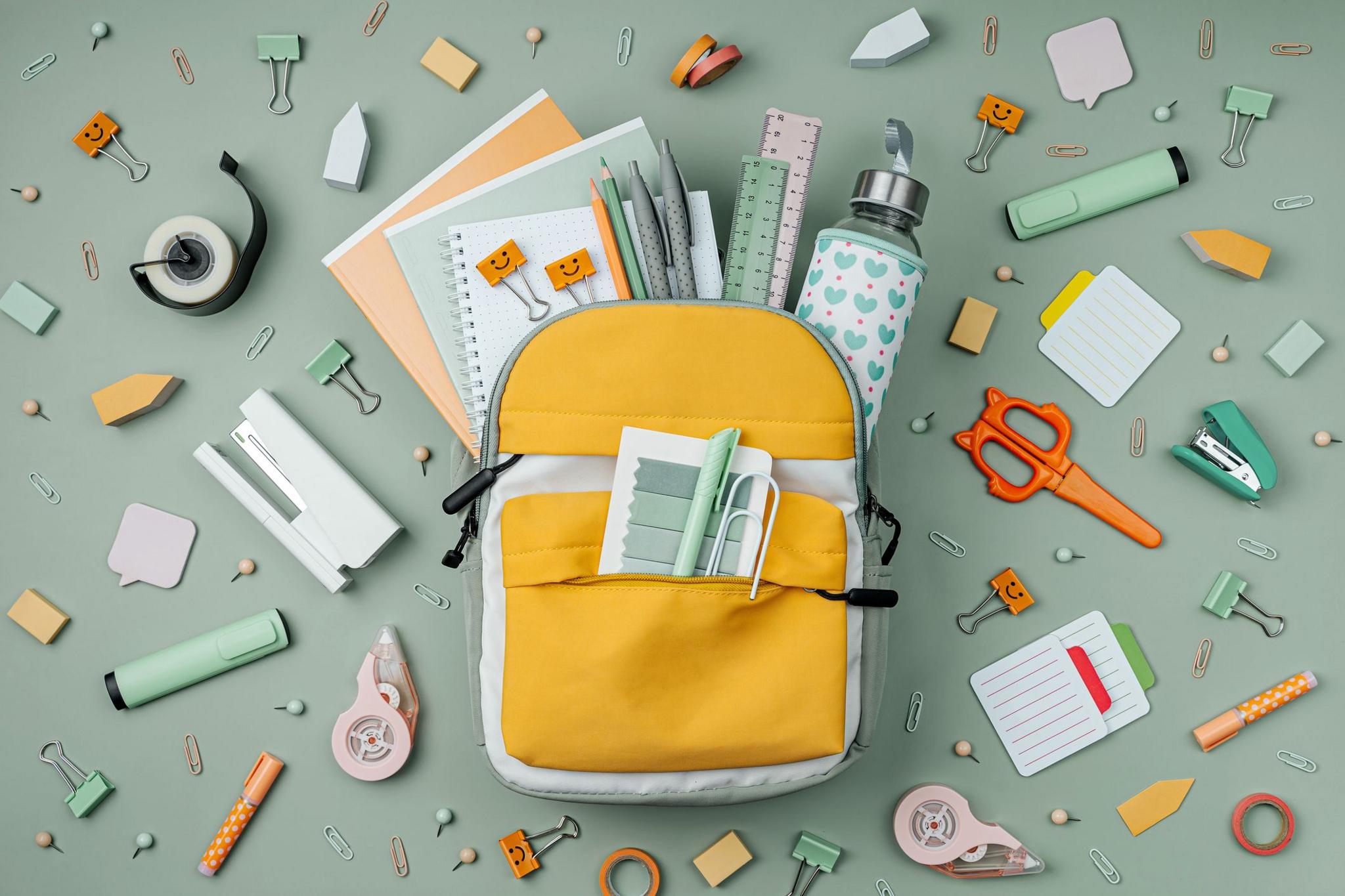 A backpack open to reveal notebooks, pens, a water bottle - the backpack is surrounded by lots of brightly coloured stationery