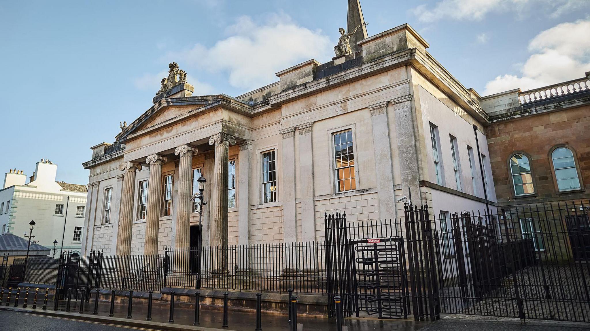 Bishop Street Court House is a large building, made of pale sandstone, with classical columns and sculptures. A large black gate surrounds the building. 