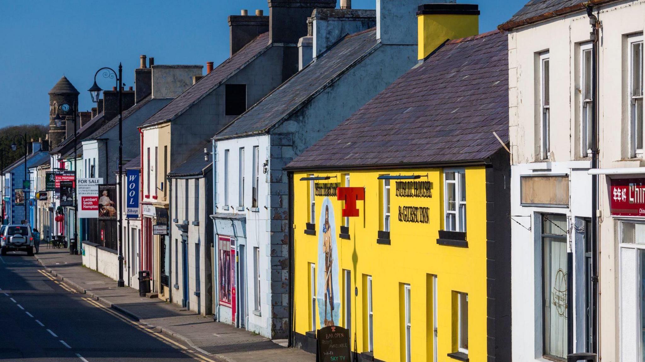 A row of buildings on a street. They include a pub painted bright yellow, alongside a number of buildings painted in pale colours. There is a round tower at the end of the street, at the left hand side of the picture.