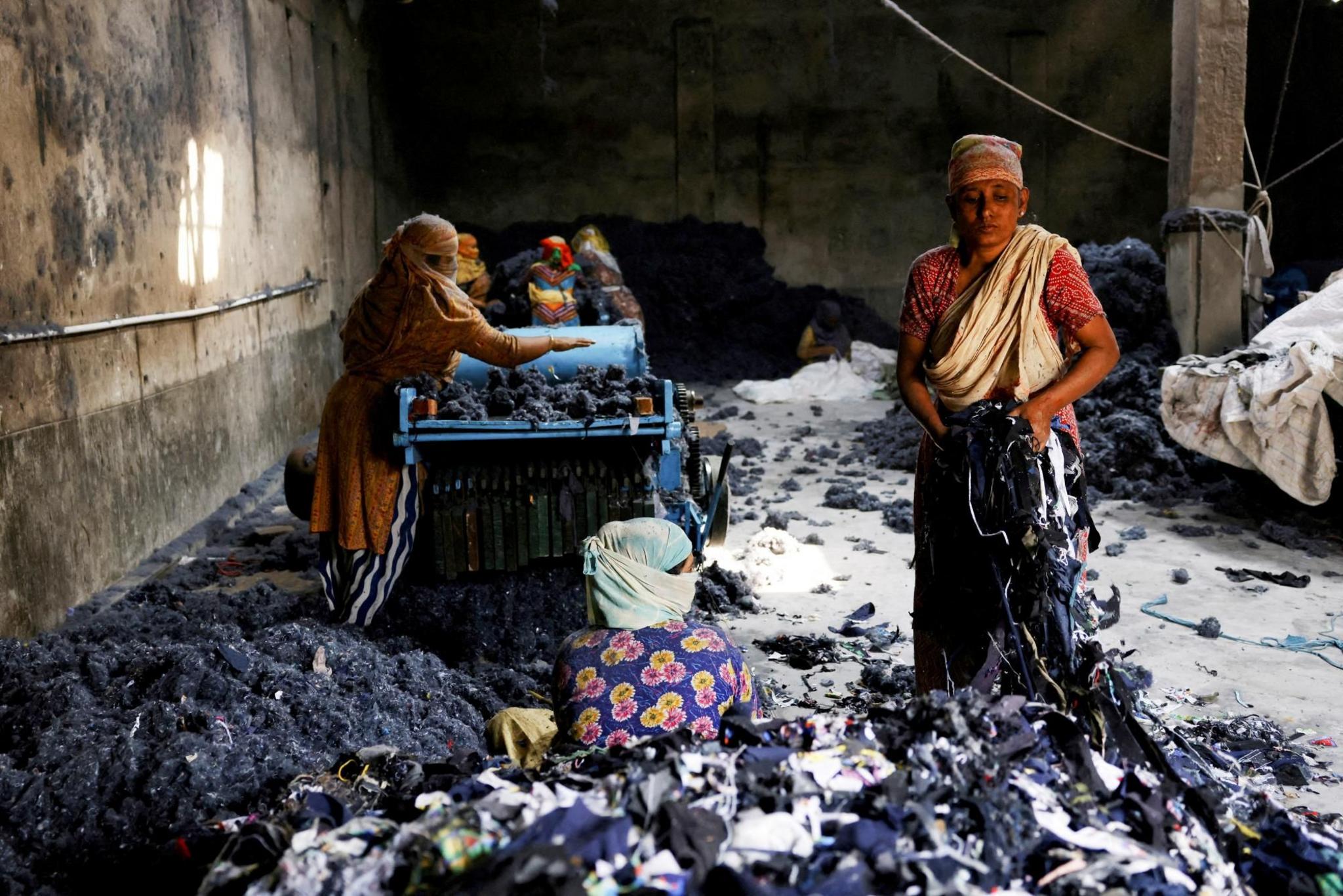 A group of Bangladeshi women gather waste clothing and run it through a machine to make mattresses. They are in a factory setting and it looks like there is black mould on the walls. One woman is sitting on the ground surrounded by material while another woman gathers it up.