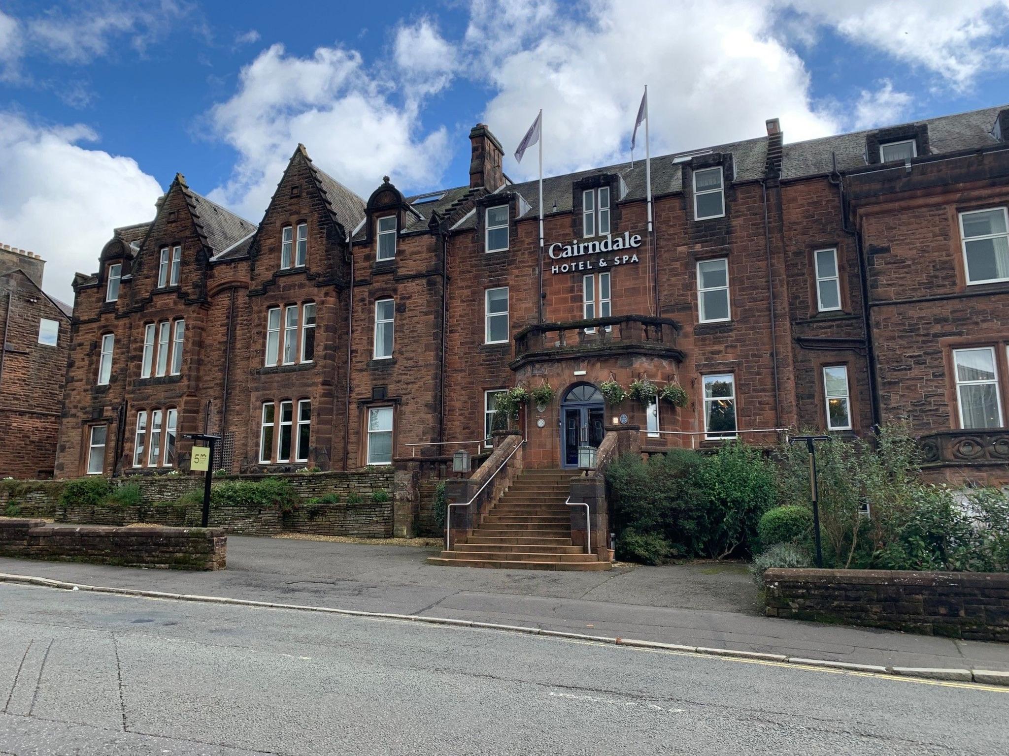 The sandstone-fronted Cairndale Hotel and Spa in Dumfries with flags flying next to a sign with its name. It has a stone staircase leading up to an arched entrance and numerous white-framed windows.