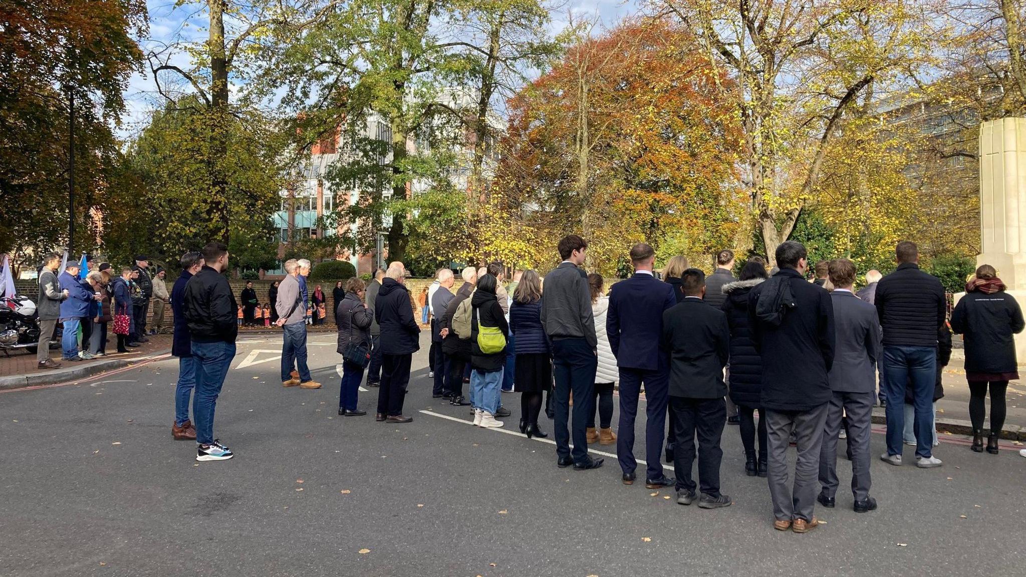 A photo of a crowd taken from behind. It's about thirty people standing in front of a war memorial with autumnal trees behind them.