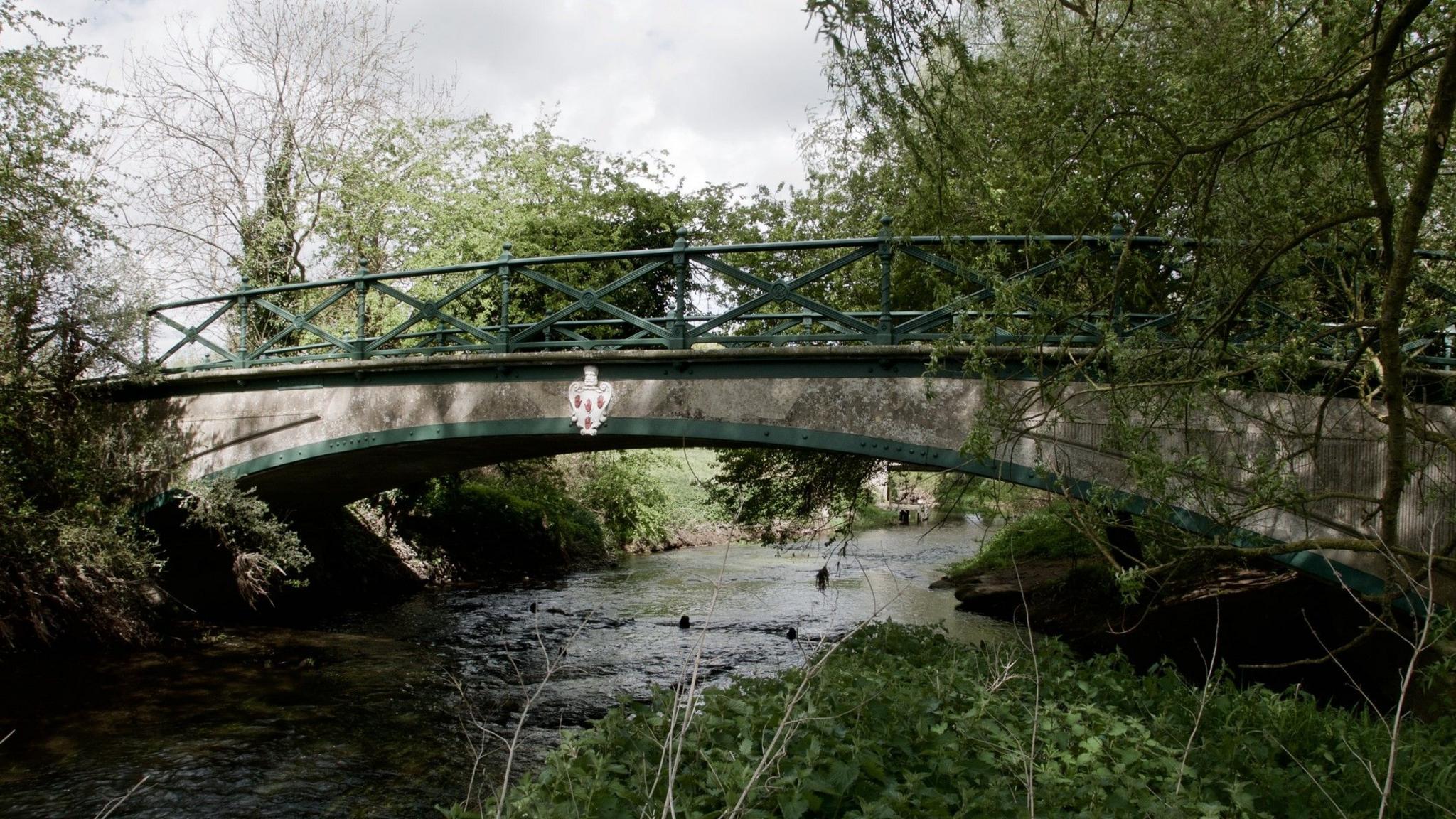 A grey bridge with green railings and arch crosses a narrow river marking the border between Norfolk and Suffolk. There is a white crest on the bridge, which is surrounded by trees.