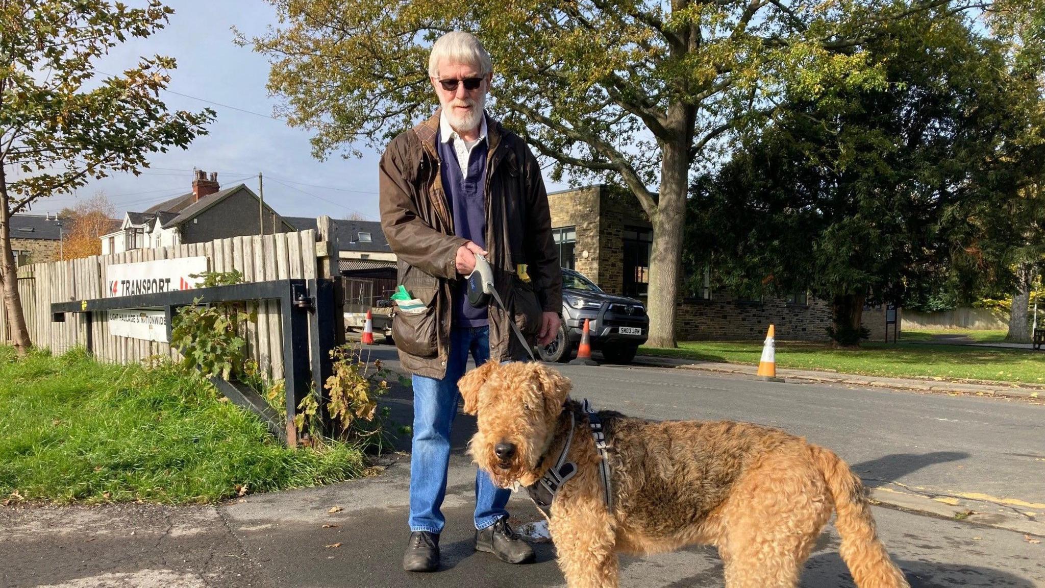 A man with white hair and a white beard is standing near a fence. He has sunglasses on and is wearing blue jeans and a dark brown coat. He is holding a lead to a sandy coloured dog of the Airedale terrier breed.