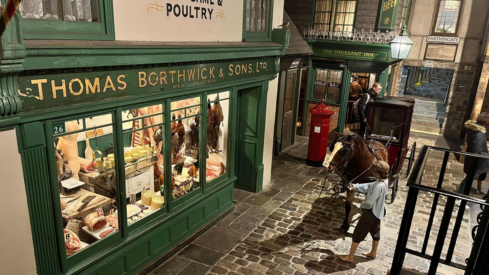 The Victorian village, with a cobbled street, a shoeless boy leading a horse through the street in front of a butchers shop