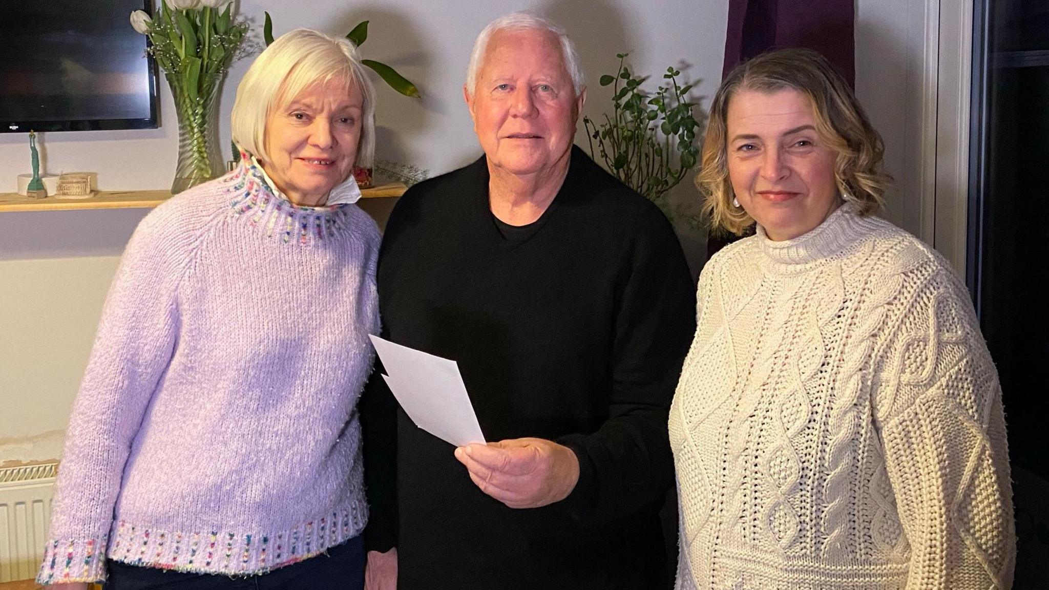 Left to right is Rowena wearing a purple jumper, Tom Phillips wearing a black jumper and Adnes Adkins wearing a white cricket-style jumper. They are looking at the camera and Tom is holding some photographs.