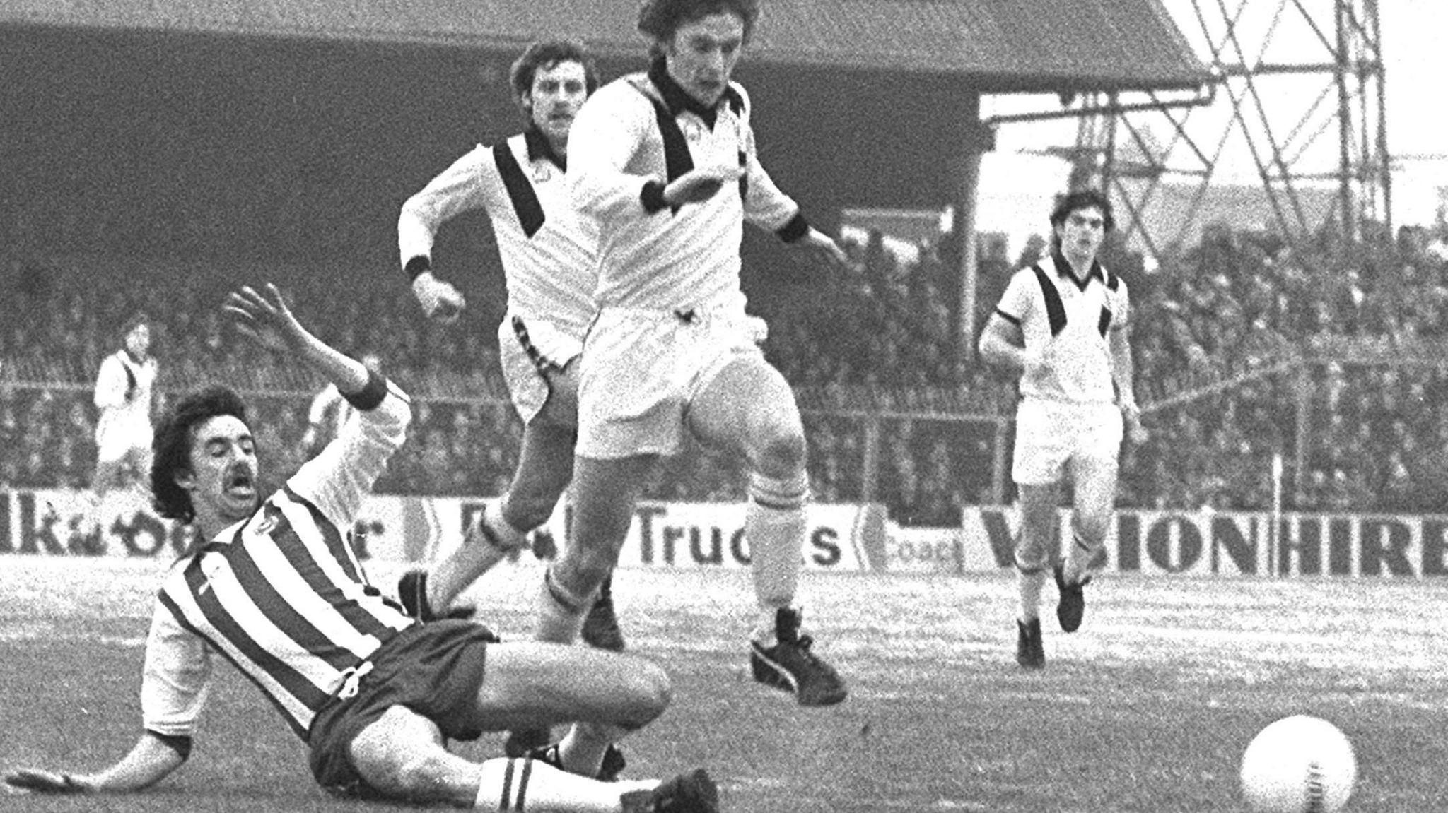 Mark Lawrenson attempts a tackle with a player running past him. A packed out crowd is in the background and the picture is black and white