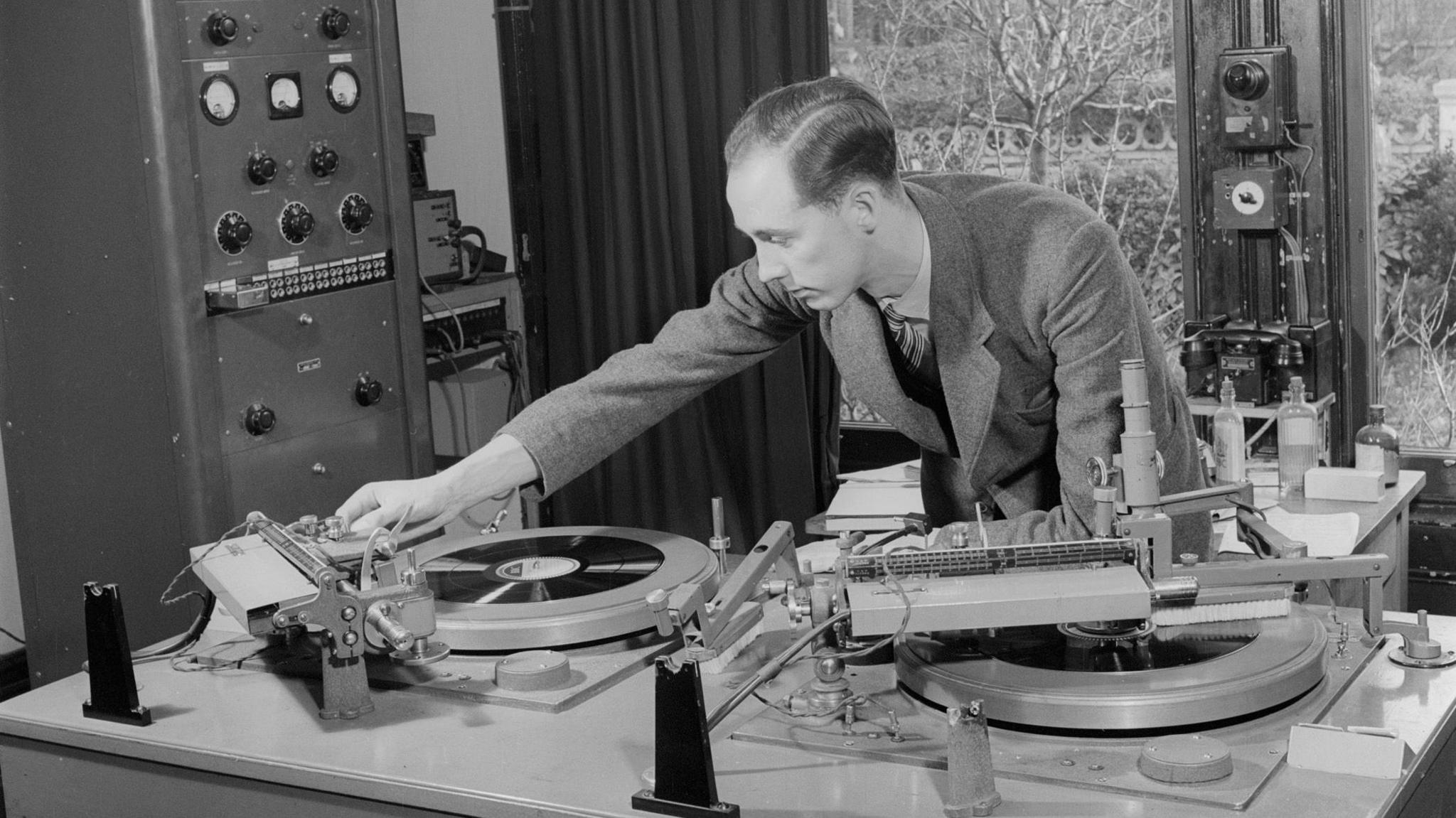 A black and white image of a man fiddling with recording equipment. There are large magnetic recording discs on the desk, and he is leaning over them to adjust the sound.