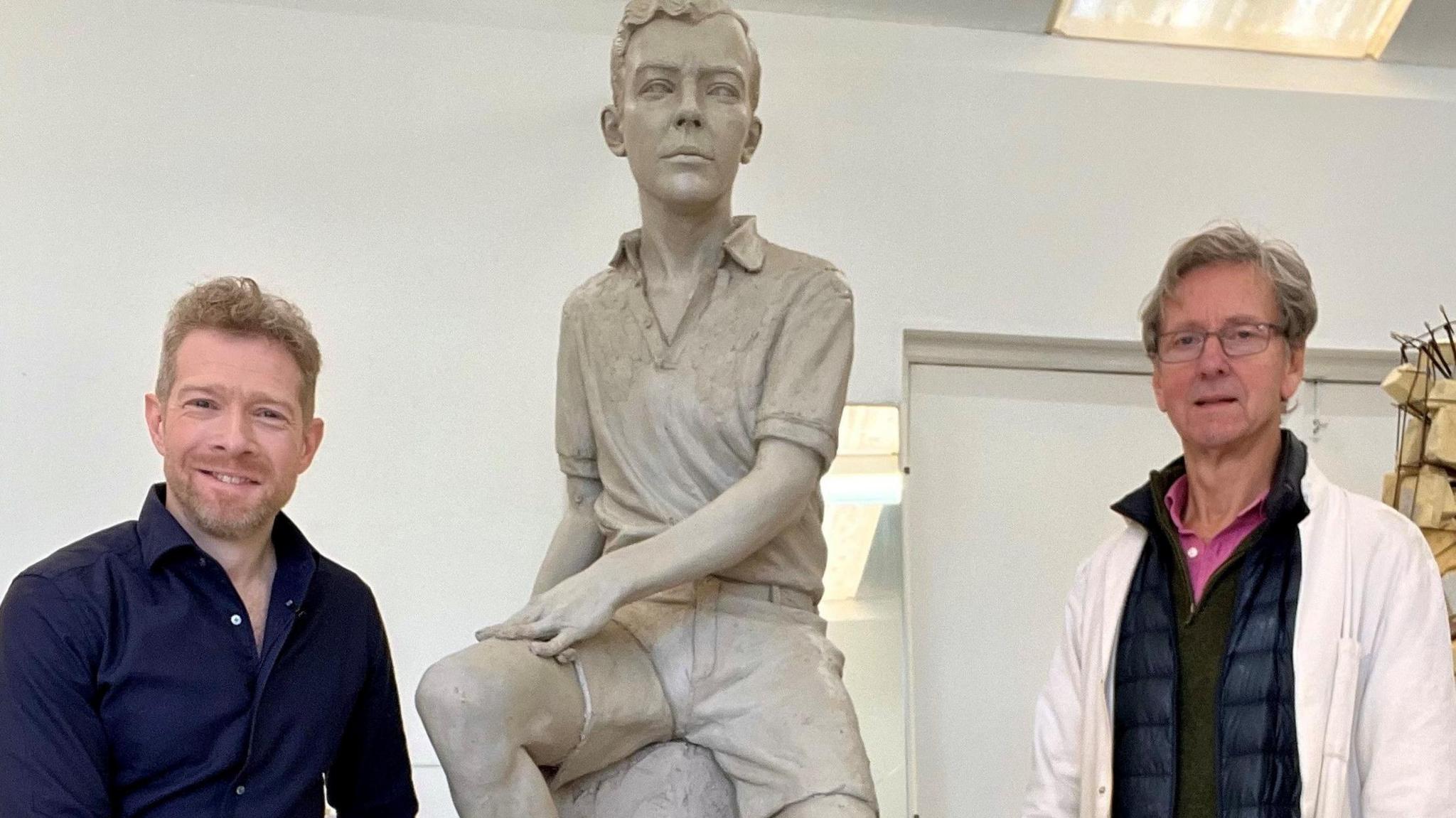 Zeb Soanes, wearing a blue shirt, and sculptor Ian Rank-Broadley, wearing a white jacket, stand either side of an alabaster sculpture of Benjamin Britten as a boy