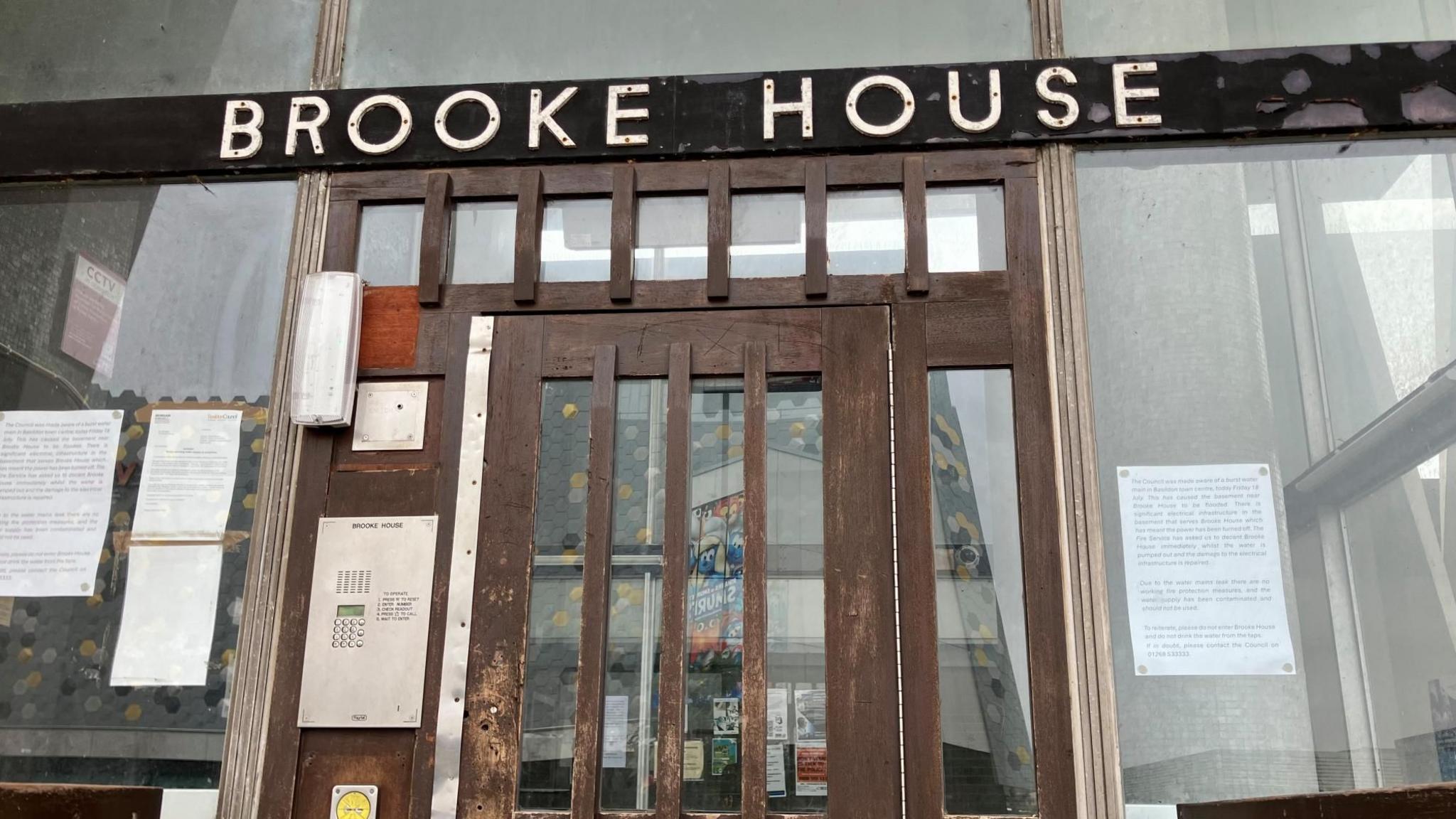 A black sign reads "BROOKE HOUSE" in white lettering. This is above a brown door with glass panels, around which multiple notices and signs are fixed. A door buzzer system is to the left of the door.