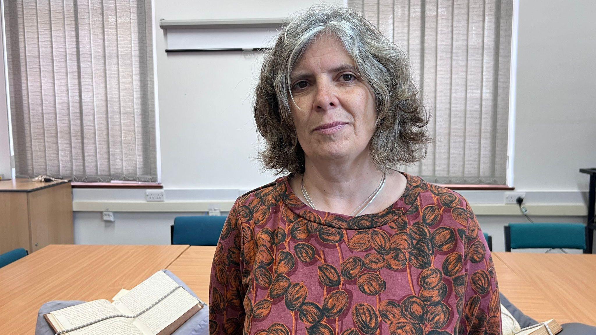 Llinos Thomas with shoulder-length grey hair stands in a room with vertical blinds that cover the windows. She is wearing a rust-coloured shirt with a dark circular pattern. Behind Llinos is a large wooden table holding two open books, each supported by angled book stands.
