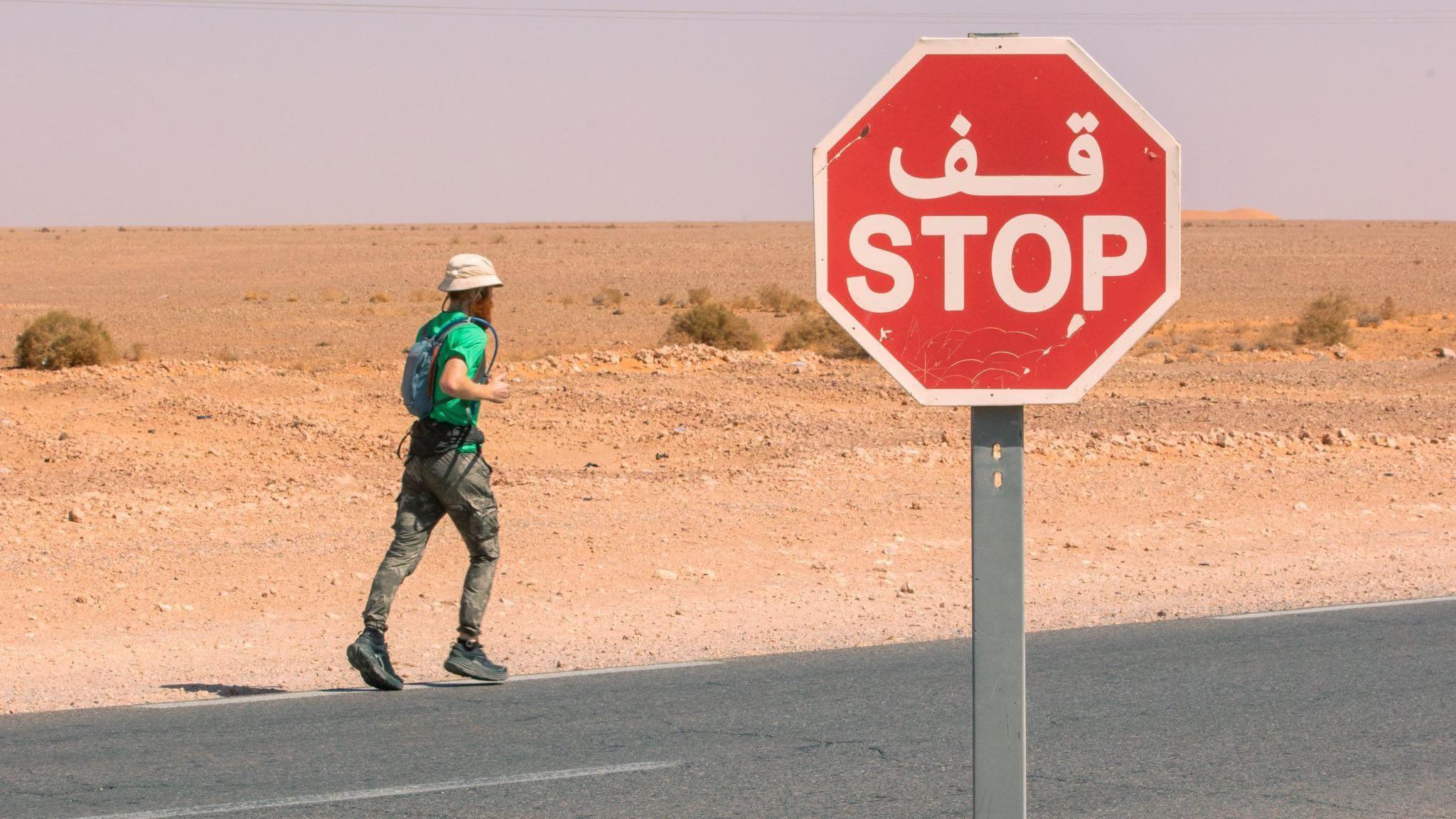 Russ Cook running along a road, surrounded by sand. He is wearing a green top and green trousers. There is a red road sign with the word  'stop' written on it. 