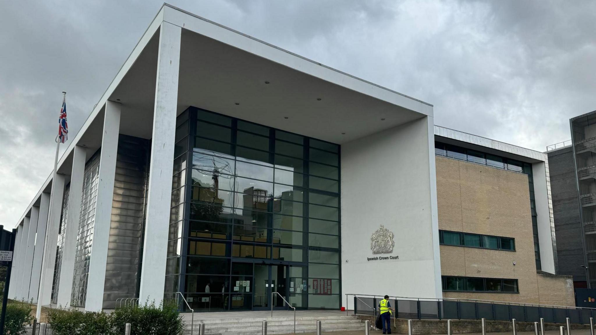 The front of the Ipswich Crown Court building. It is a cloudy day. A person in a high-vis yellow jacket appears to be tidying up outside the court entrance.
