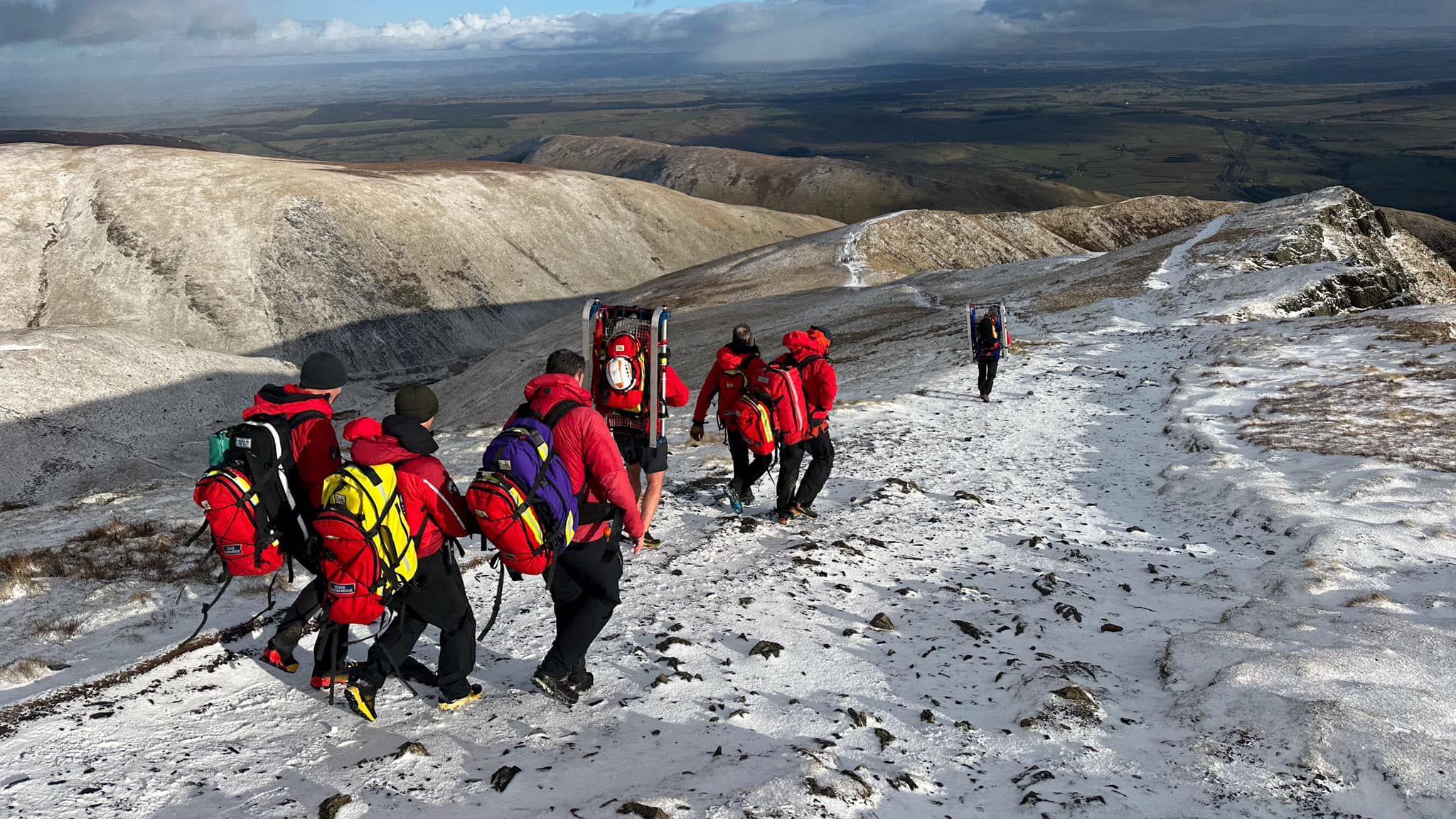 Keswick Mountain Rescue volunteers, who are all wearing red and carrying large backpacks, a stretcher and rescue gear. They are descending from Blencathra which is covered in snow with parts of the rock showing through. 