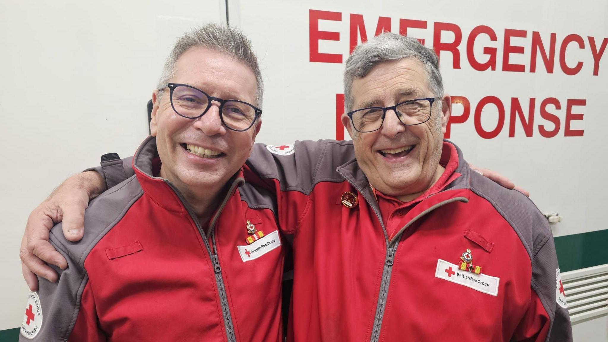Andrew and Steve are smiling at the camera with their arms around one another. they both have short grey hear and are wearing black rimmed glasses. They are wearing matching red cross jackets. behind them is a vehicle with the words emergency response on.