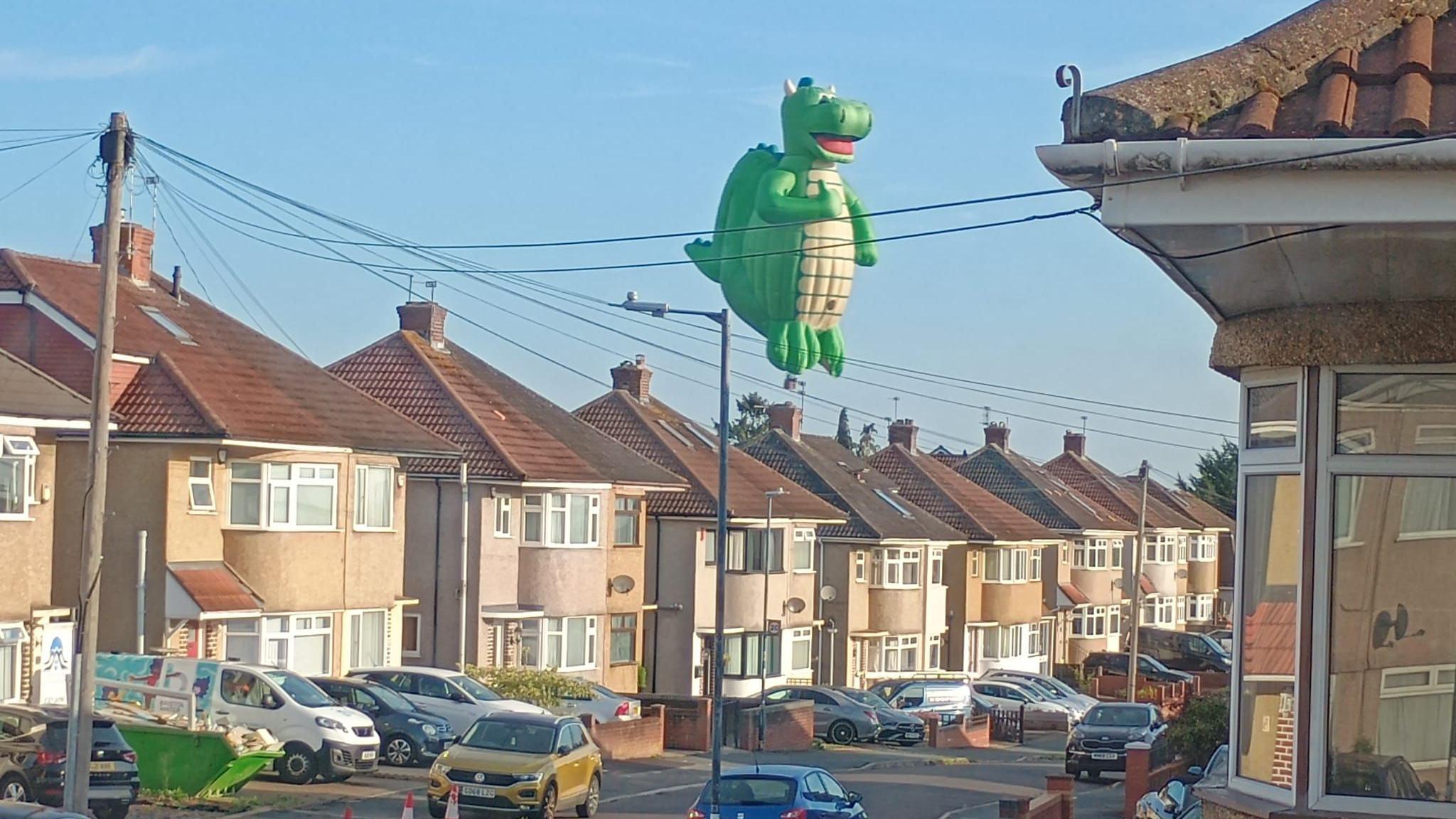 A large dragon-shaped hot air balloon is flying over residential streets with cars lined up.