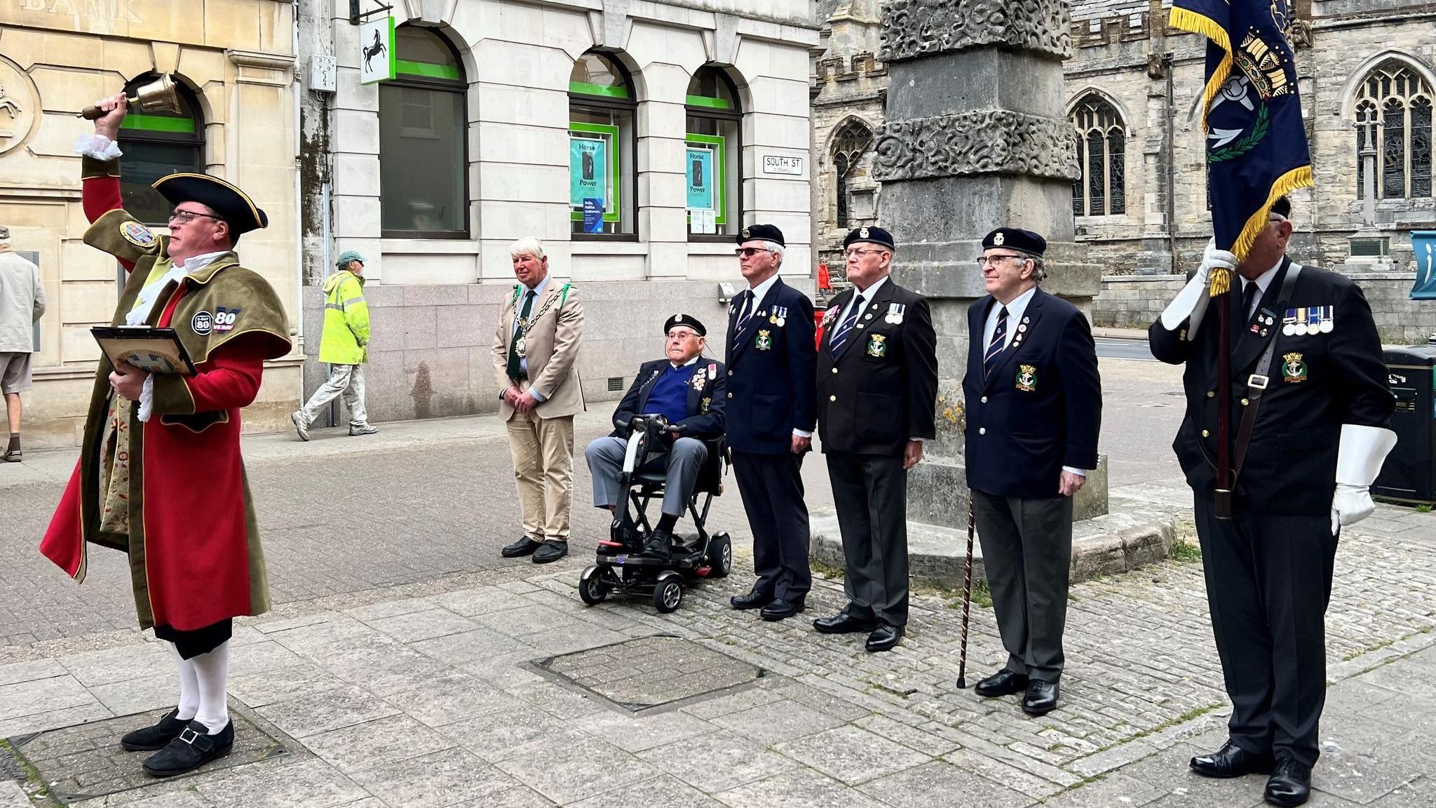 Five Royal Navy veterans and the mayor lined up in front of a stone monolith. In front of them is the town crier in his traditional red coat and tricorn hat, ringing a hand bell.