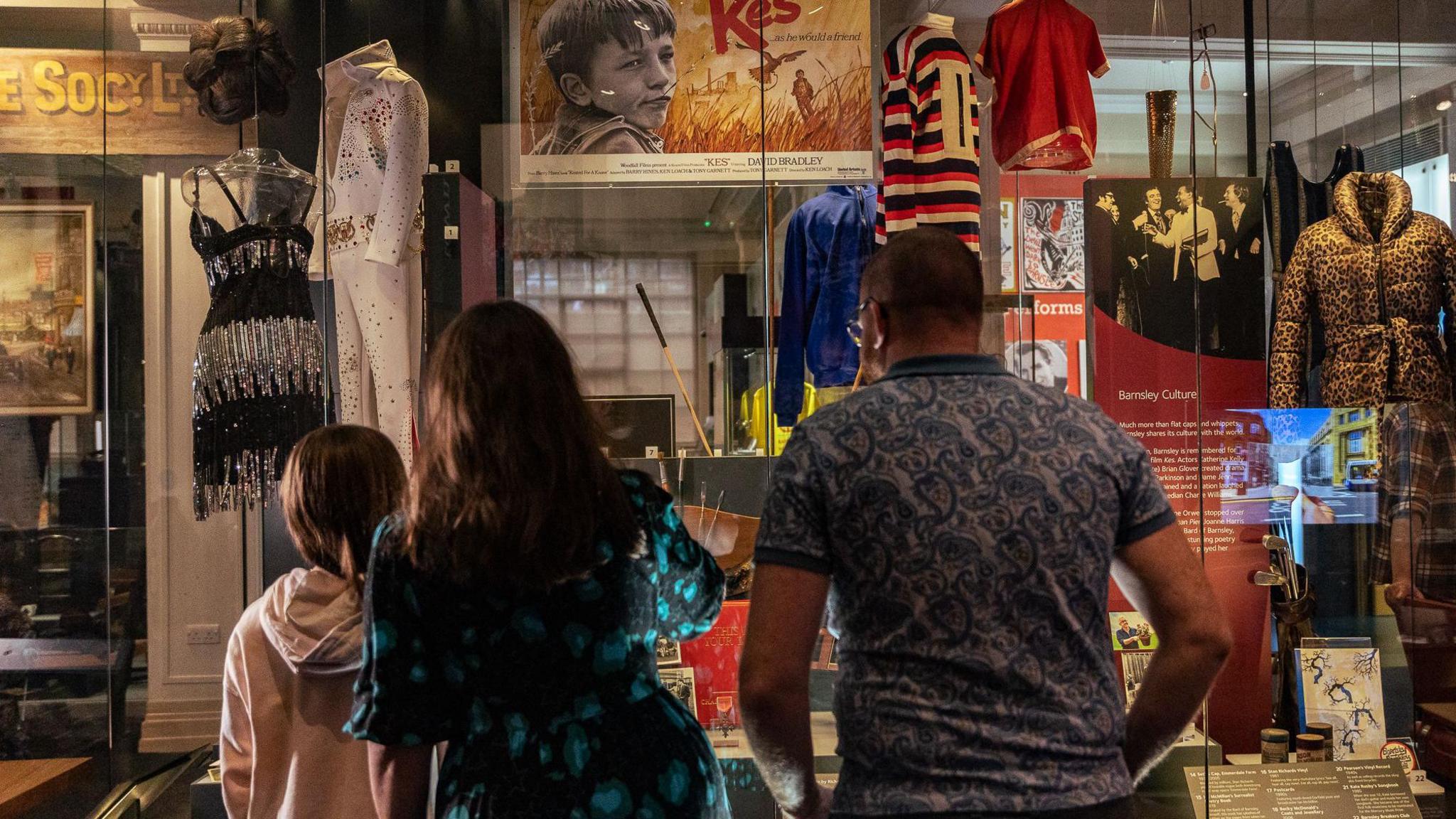 A girl, aged about 10, in a pink hoodie looks into a glass case at a museum. Next to her are her parents, also both facing away from the camera and into the cabinet. Her dad wears a blue paisley polo shirt and mum wears a black and turquoise patterned summer dress. Lots of outfits on mannequins, some movie posters and information notices are inside the cabinet.