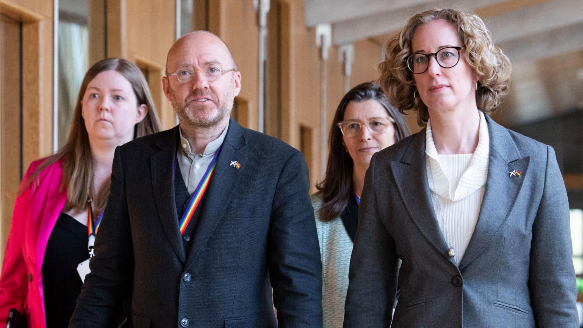 Scottish Green Party co-leaders Patrick Harvie (second left) and Lorna Slater (right) walking in the Scottish Parliament, with two women walking behind them. Mr Harvie is wearing a three-piece blue suit and an open necked shirt, while Ms Slater is wearing a grey suit jacket and a cream top.