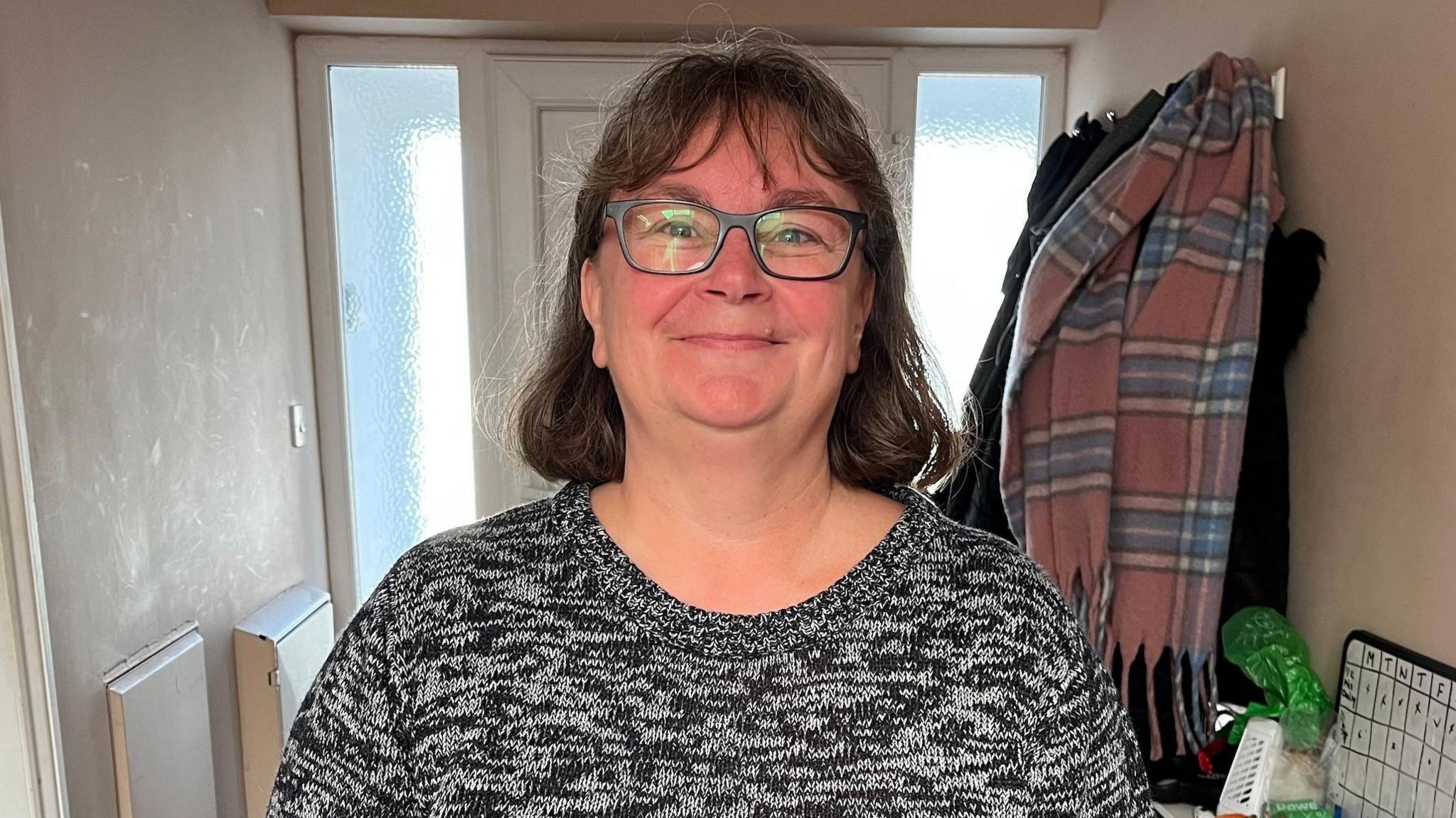 A woman with glasses and a wool jumper is smiling to the camera. She is standing in the hall of her home.