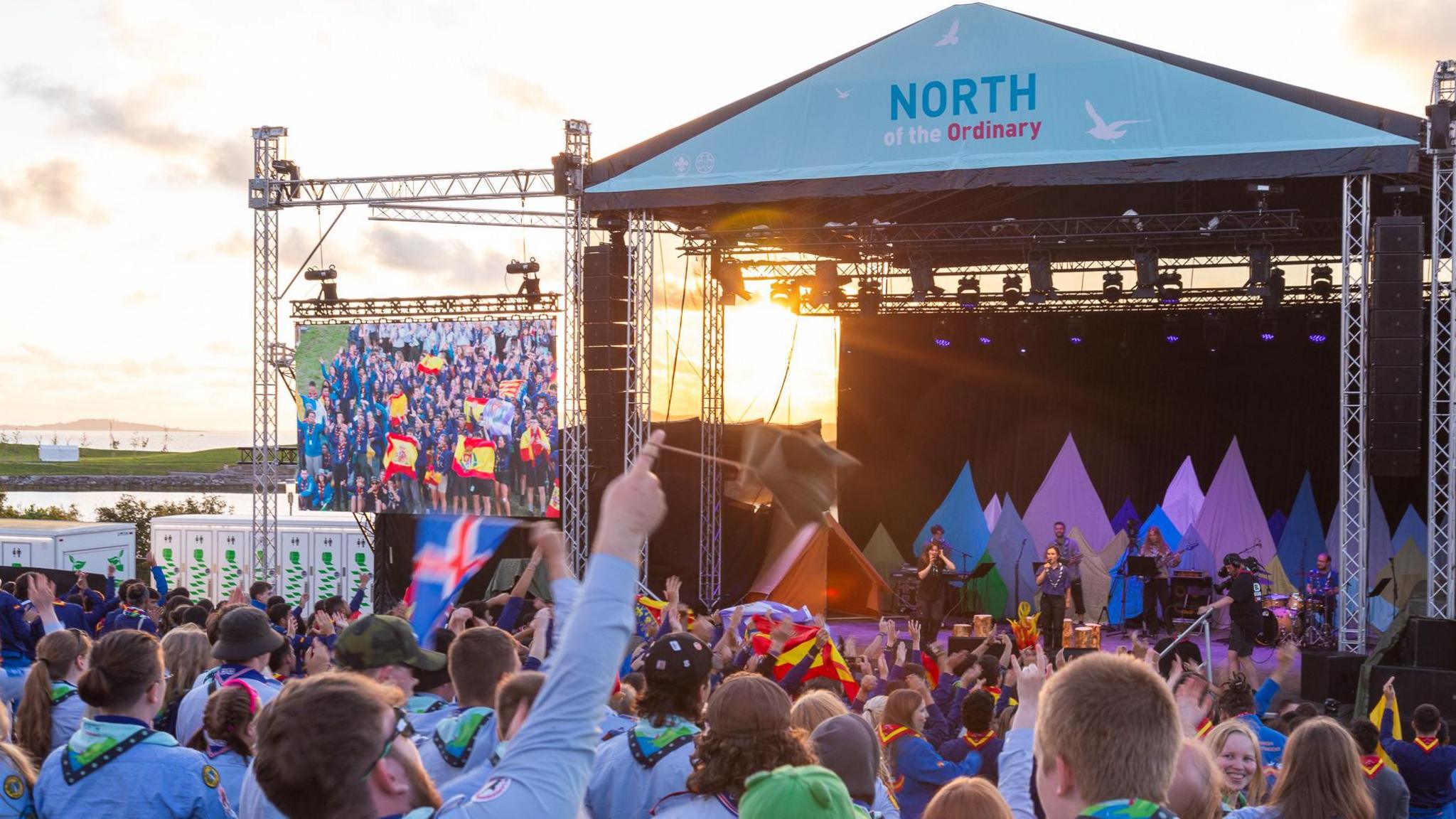 Scouts in uniform look towards a stage. Many have flags.