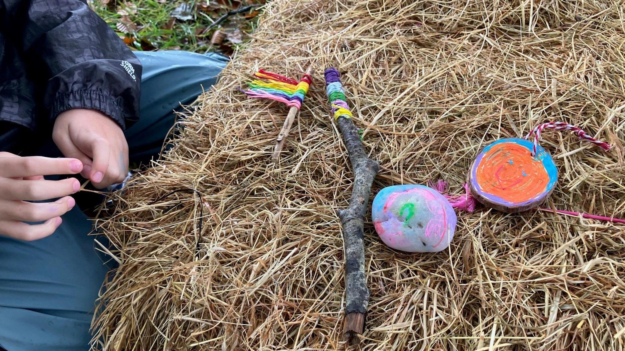 Brightly painted pebbles and twigs with coloured wool tied on have been placed on a hay bale. A child's hands are visible on the left. There is grass in the background.