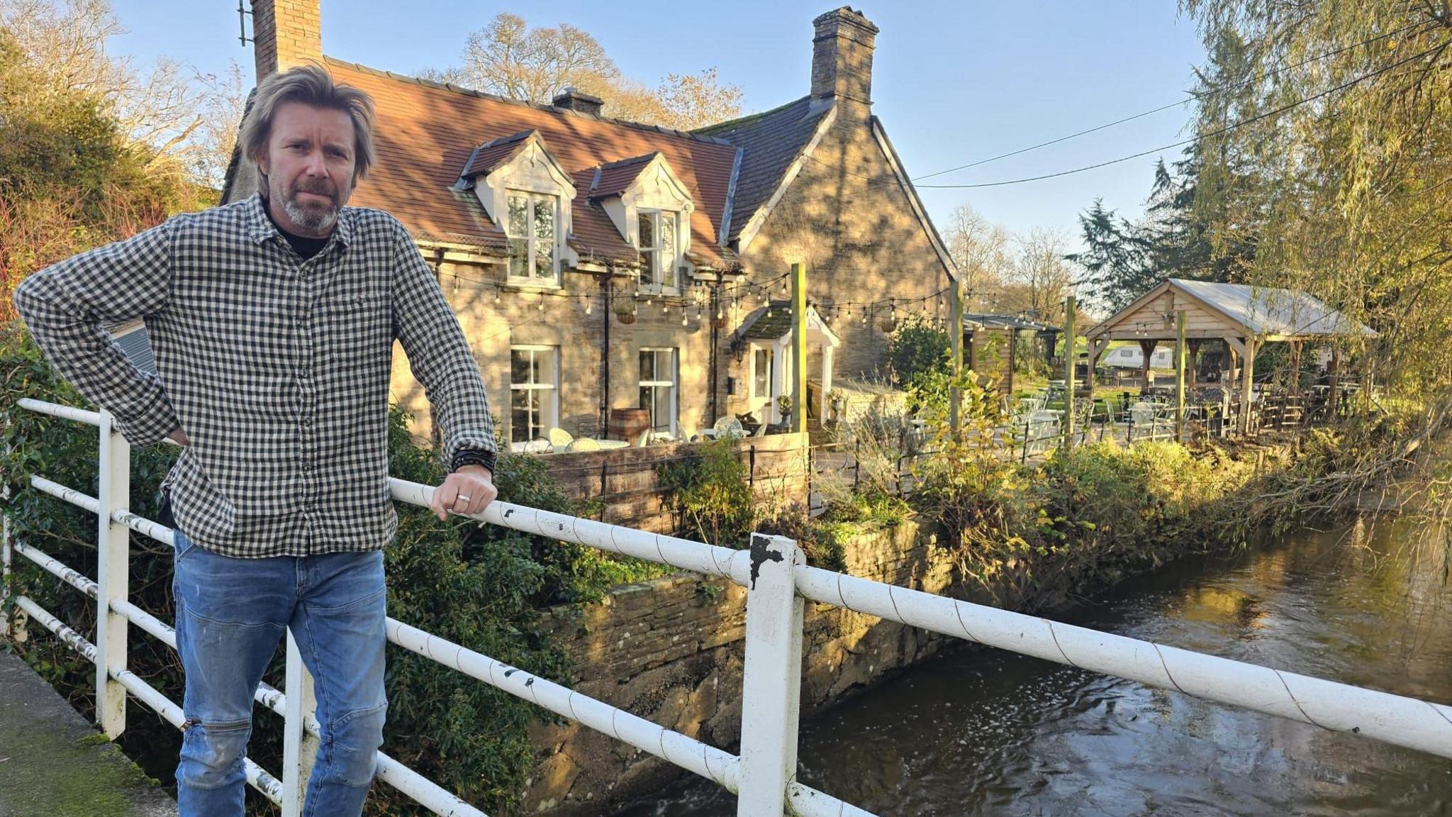 A man with swept back blond hair in a black and white checked shirt and blue jeans leans on a white railing with a country pub behind him and a river running alongside it. 