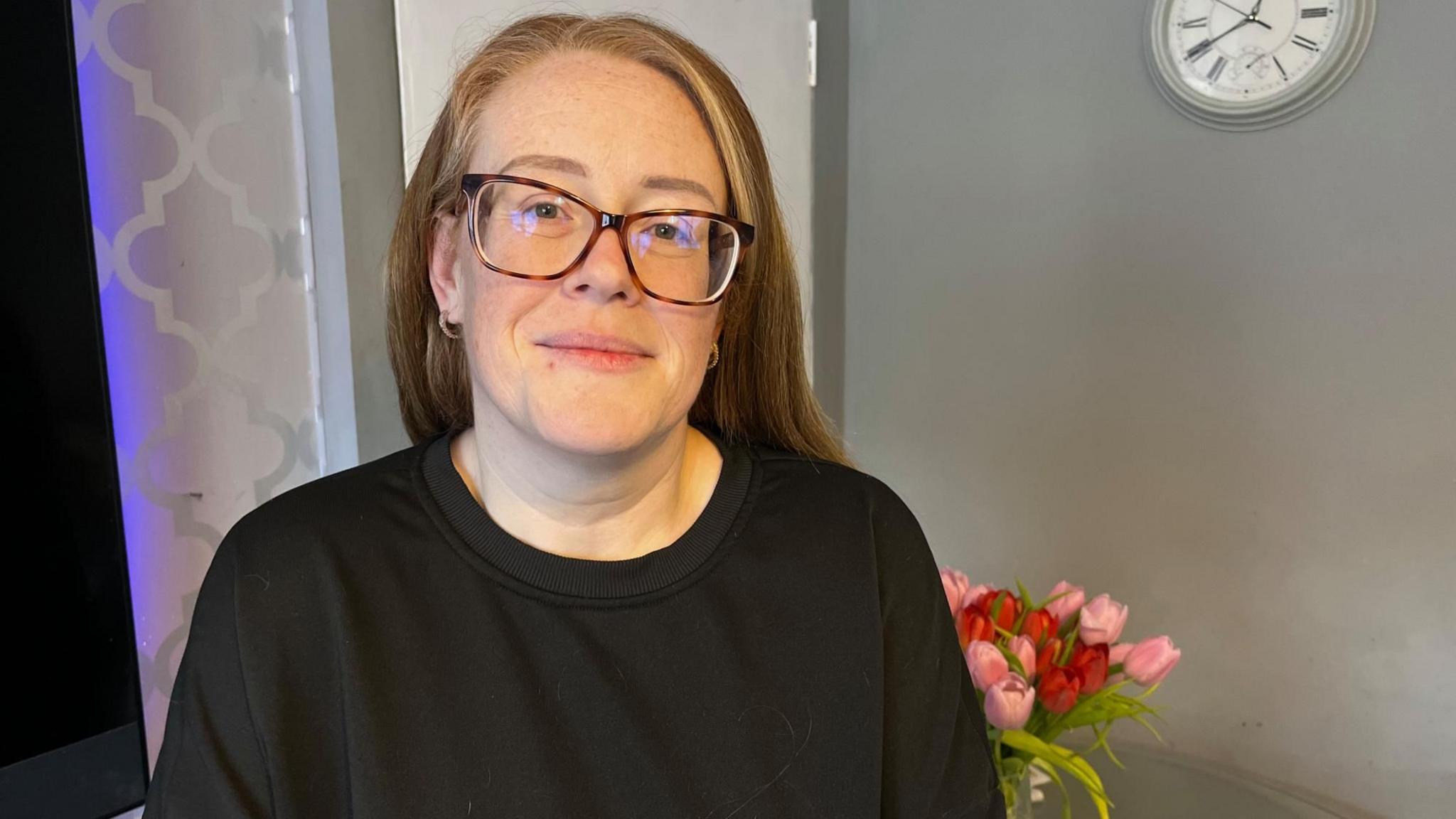 Lynsey Boyd  smiles in a grey room with graphic wallpaper, a vase of pink and red tulips in the background. She is wearing a black top and has tortoiseshell glasses on