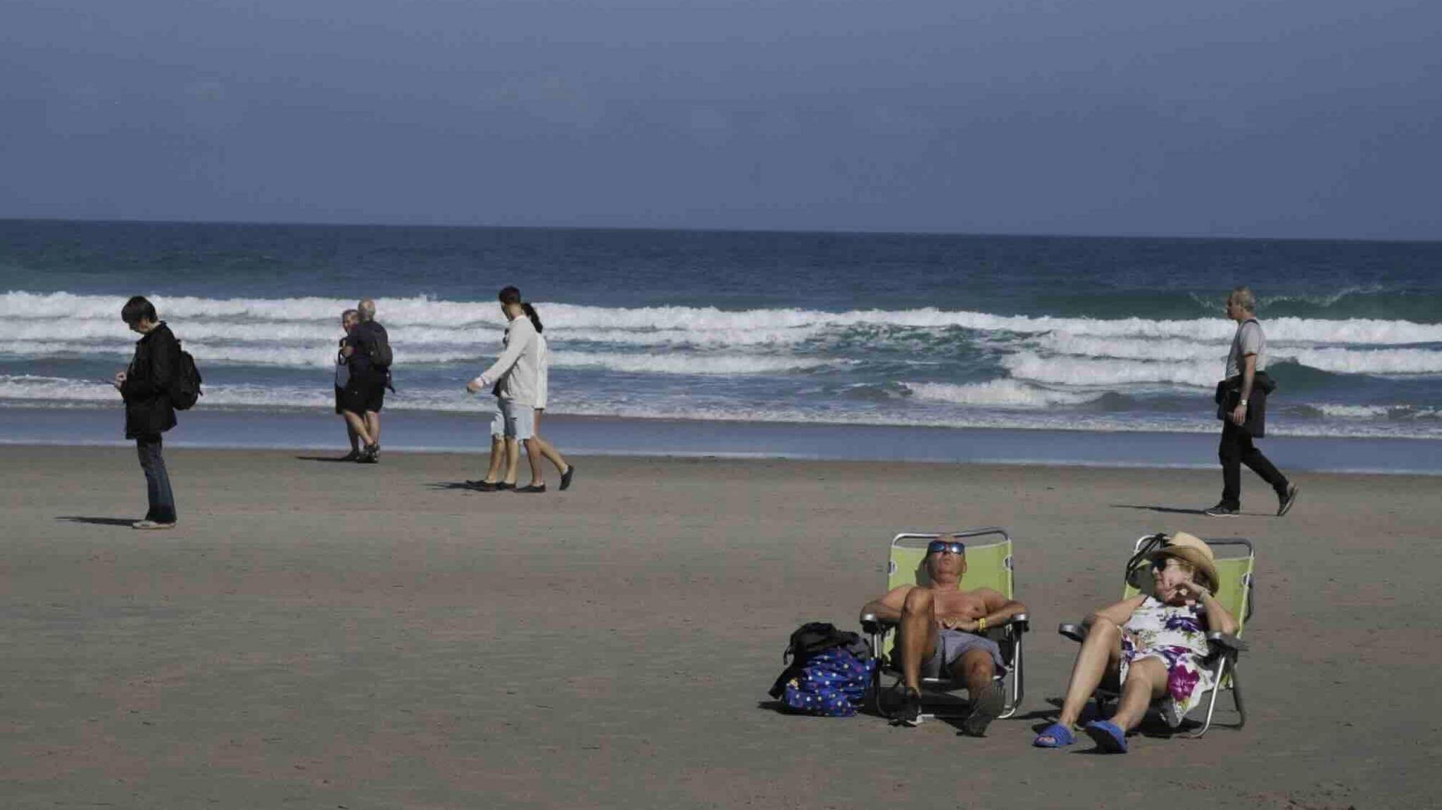 A man and a woman lying on a chair on a beach with people walking and waves breaking behind them