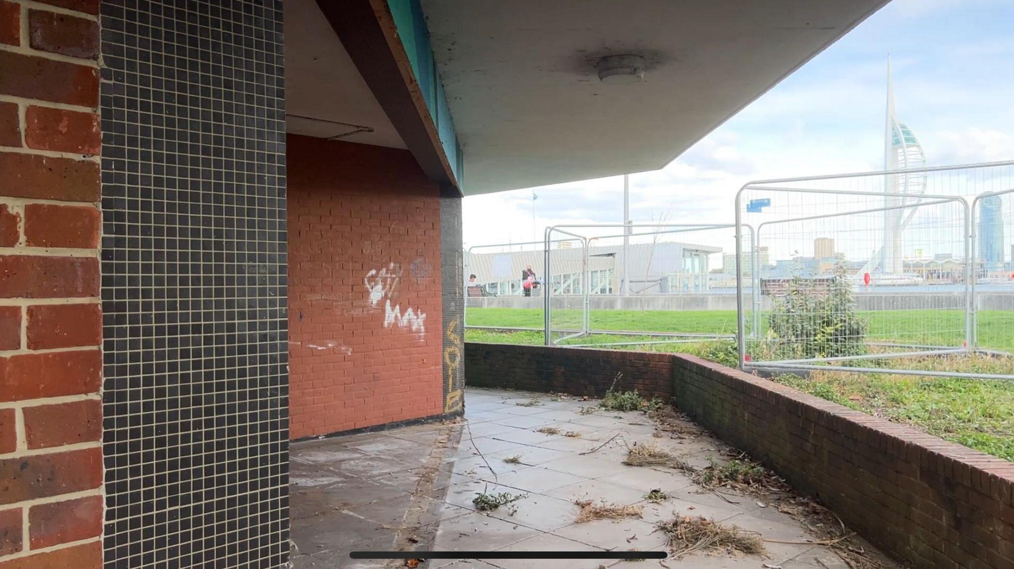 To the left a brick and tiled wall of the old bus station with graffiti on and to the right, you can see through the fencing to the water and Spinnaker tower.