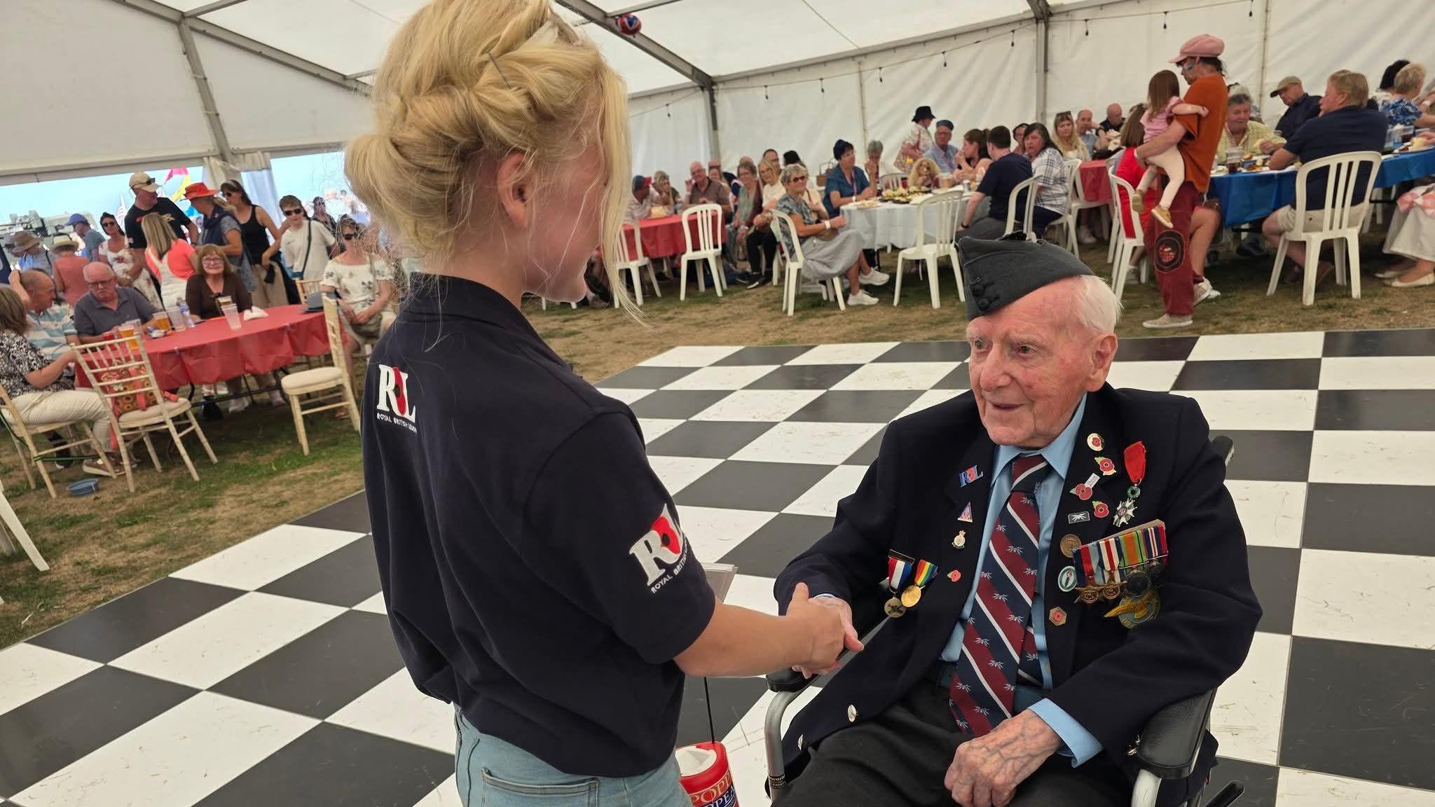 A young girl with blonde hair is wearing a dark blue T-shirt that has the Royal British Legion logo on it with a red poppy. She is shaking the hand of an elderly veteran in a wheelchair. He is wearing a dark green army hat, a blue shirt, a red and navy tie, and a black suit jacket that is adorned with badges and medals. There are people sitting on white chairs in the background.