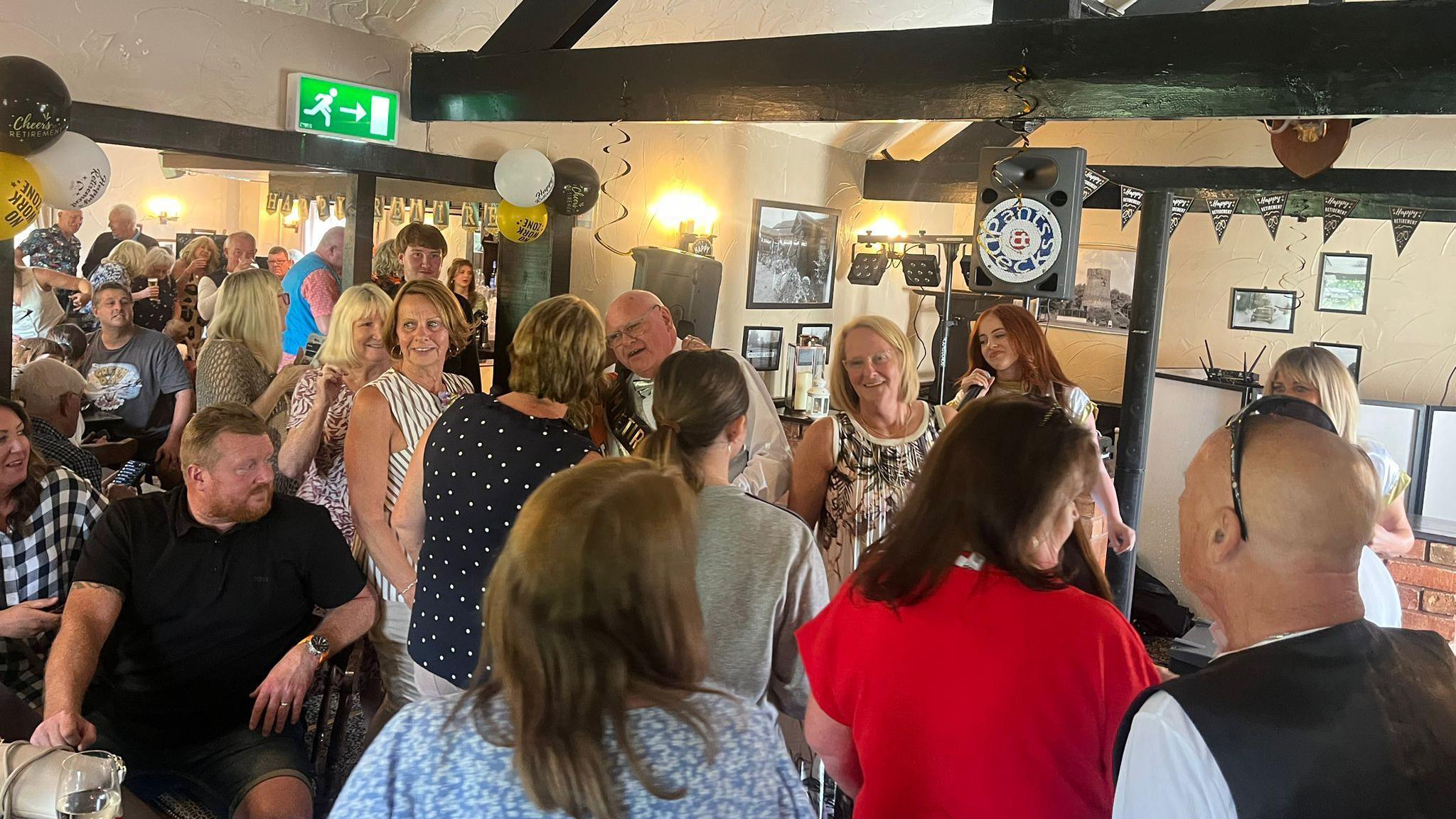 Dozens of people gather in a pub for a party. An auburn haired woman is singing into a microphone