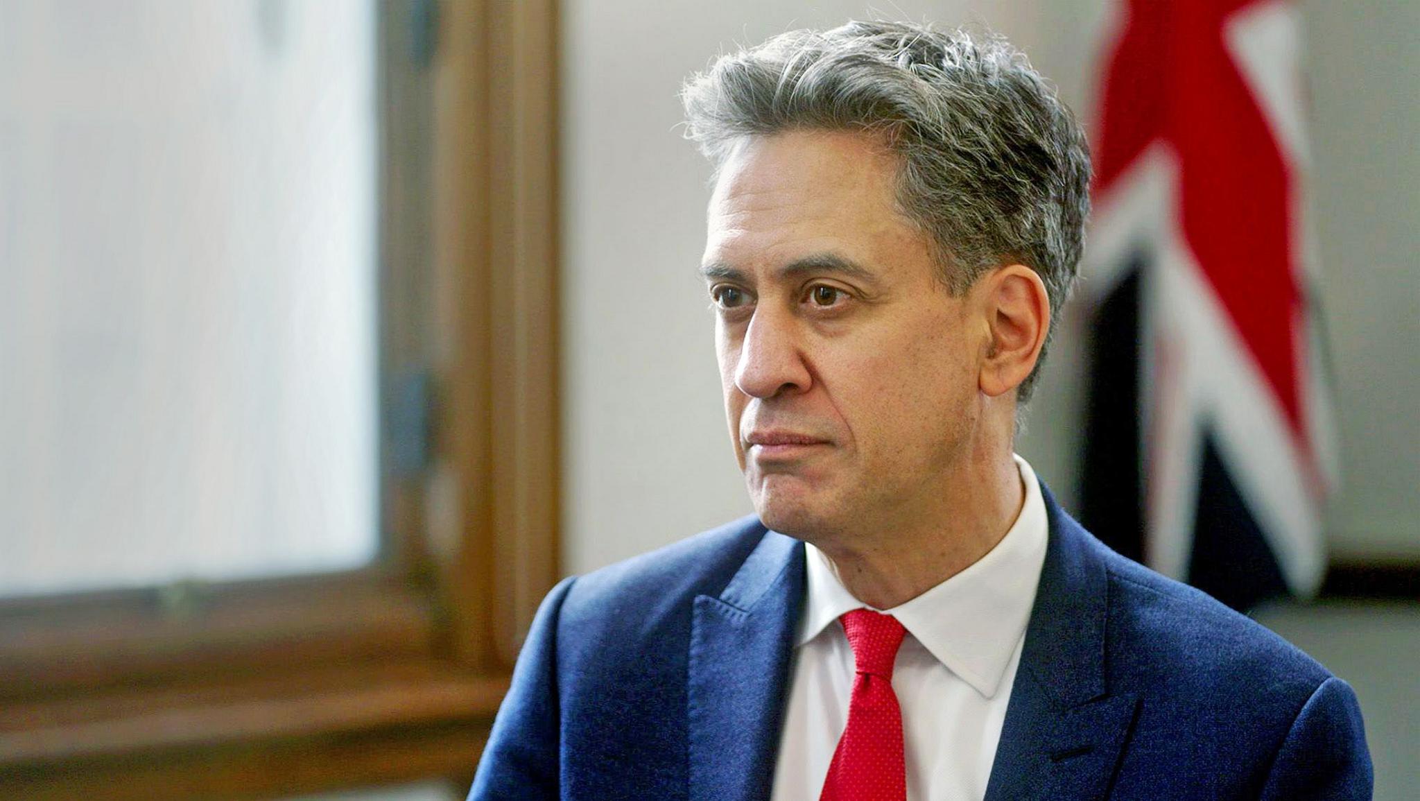 Ed Miliband pictured during his interview. He has short, dark, greying hair. He is wearing a navy suit, white shirt and red tie. Behind him is a wooden window frame and a Union Jack flag. 