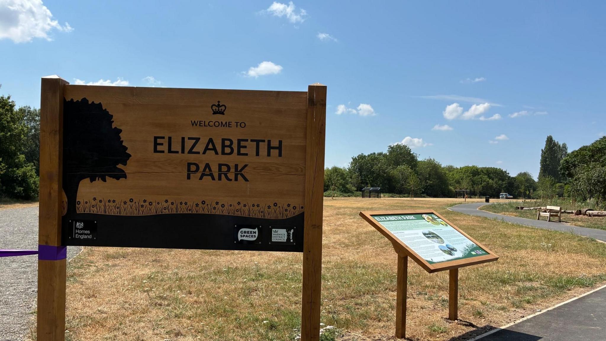 A wooden sign with the words Elizabeth Park stands in a grass field. The grass is brown and dry from the summer heat. Next to it is another wooden sign with a map of the park. A gravel path and a tarmac path are either side of the signs.