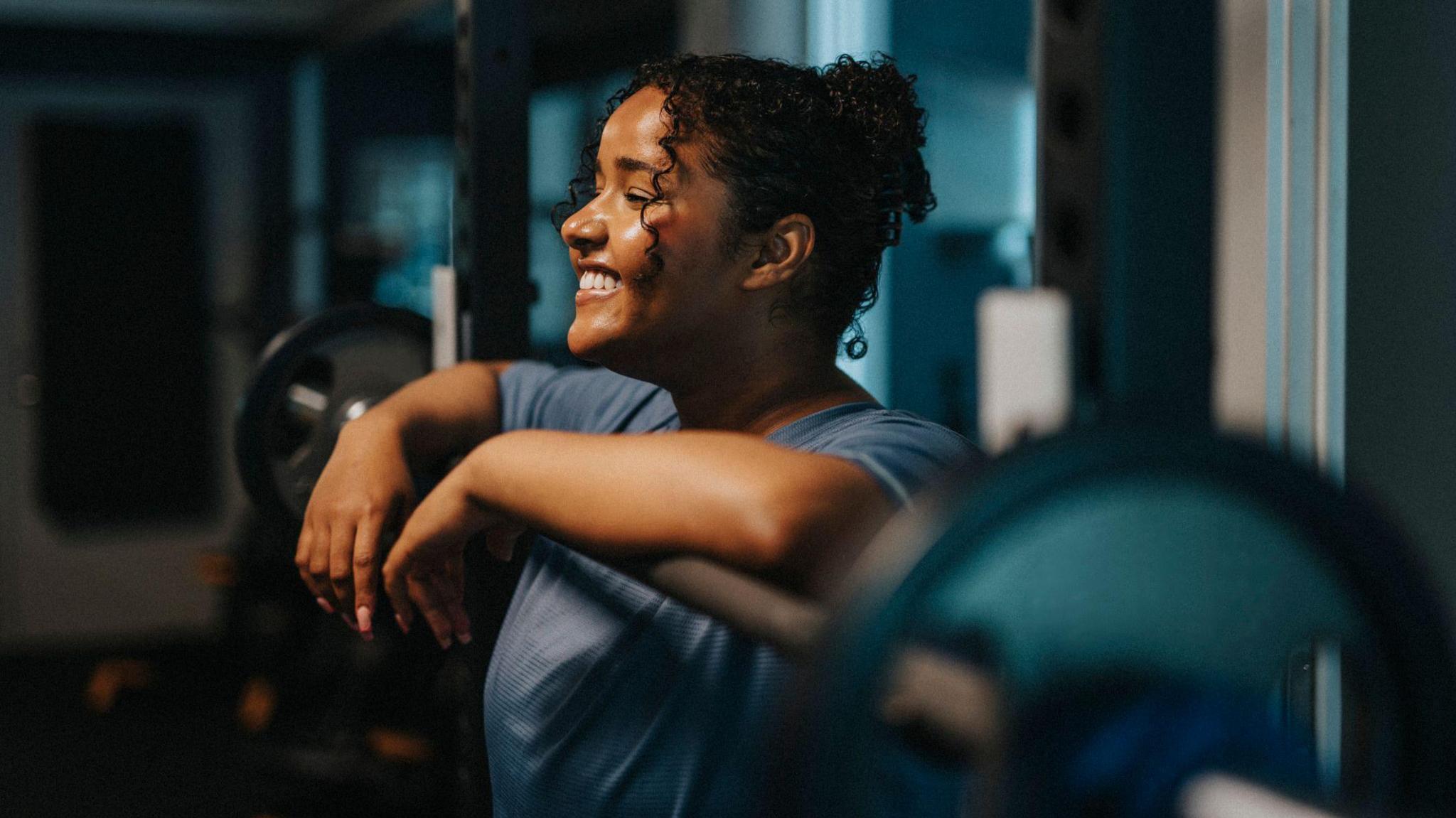 A stock image of a women in a t-shirt with her forearms draped over a barbell on a squat rack.