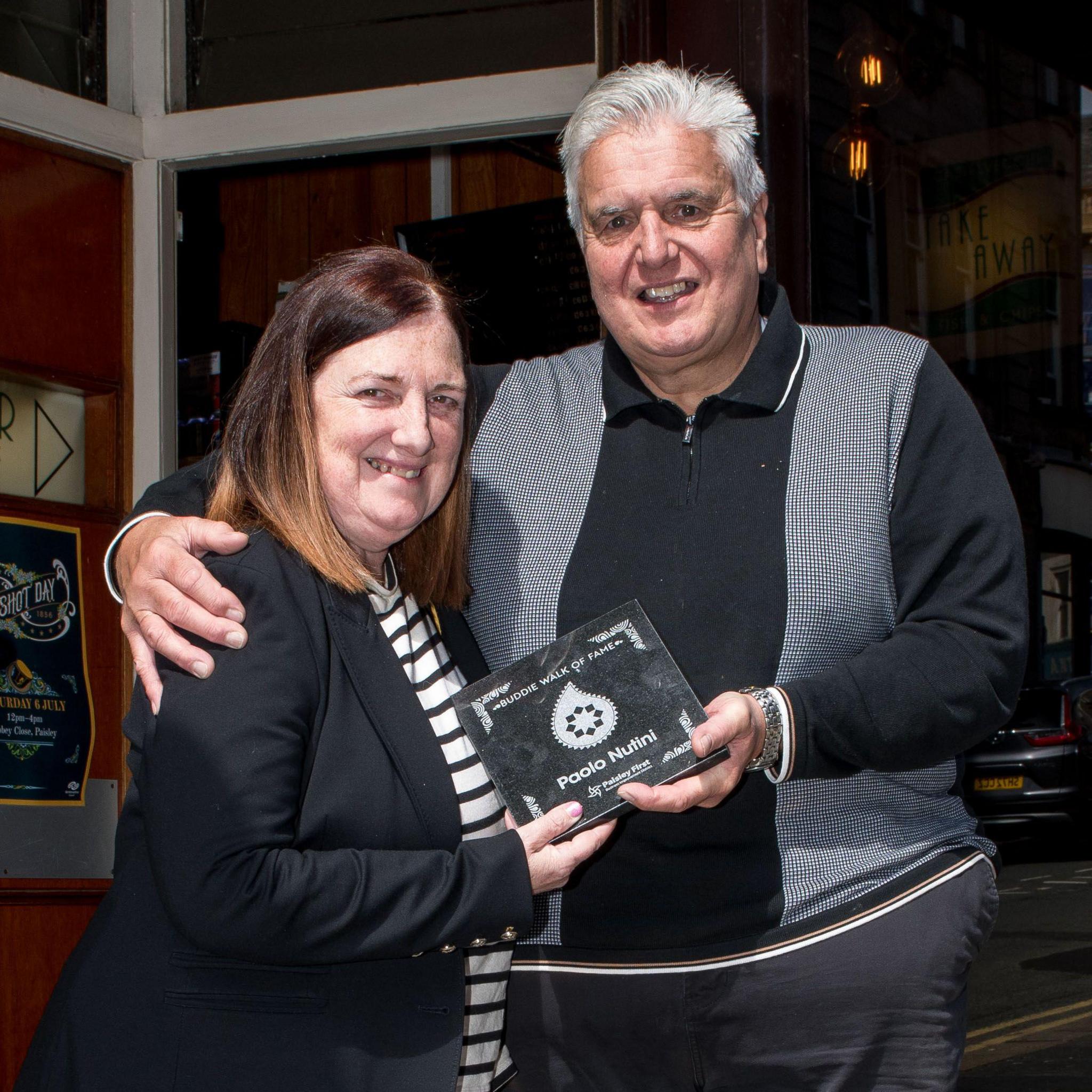 Alfredo and Linda Nutini holding a small black award with a white paisley pattern on it that reads in white "buddie hall of fame, paolo nutini".