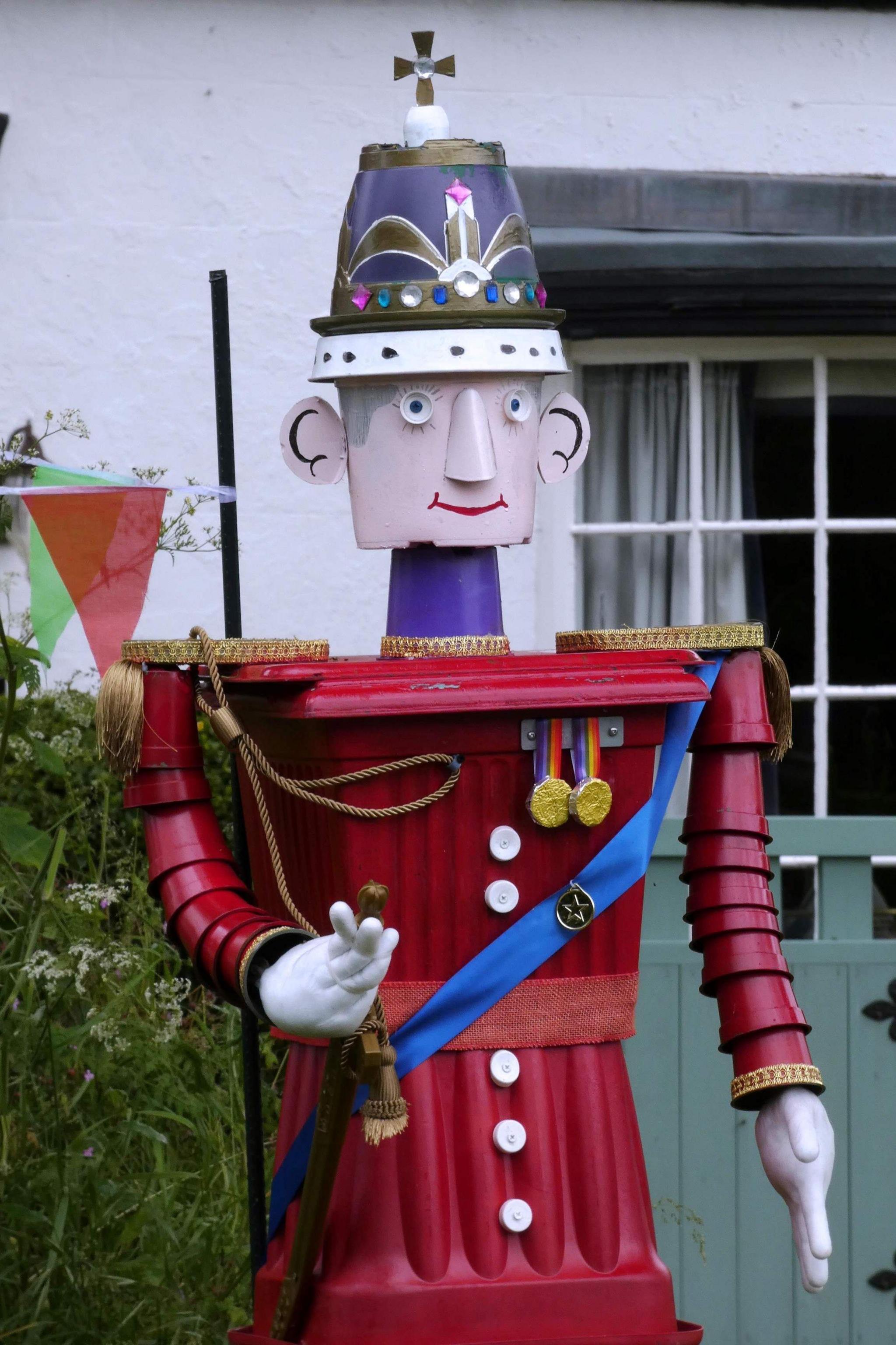 A model of a man made from plant pots in a red uniform, medals and a cross on his hat. There is a building behind it with a window, a gate and bush.