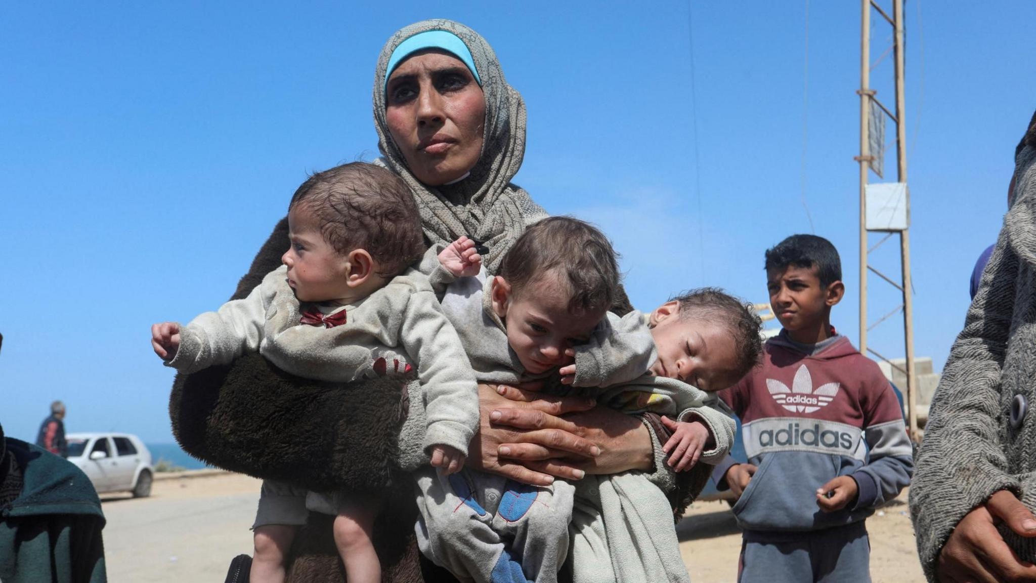 A Palestinian woman carries three babies in central Gaza after fleeing al-Shifa hospital in Gaza City, on 21 March 2024