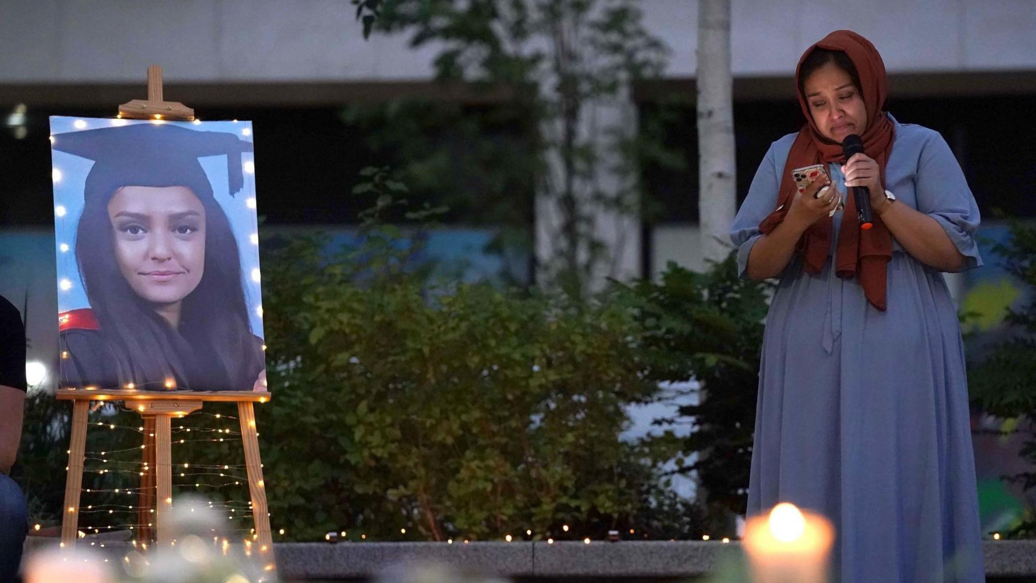 Jebina Yasmin Islam reads from a phone at a vigil for her sister, the murdered teacher Sabina Nessa. A lit candle is blurred in the foreground. A graduation photo of Sabina is on an easel, surrounded by small, white fairy lights