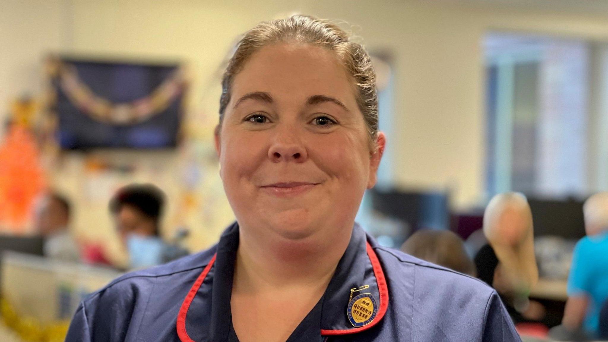 Katie Andrew wearing a blue NHS uniform with two lanyards, her hair tied back in a bun, she smiles at the camera