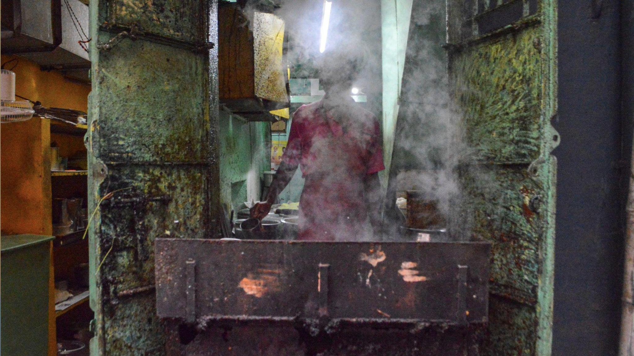 A man in a pink shirt cooks food on a hot open stove, flanked by grimy steel doors, shrouded in steam