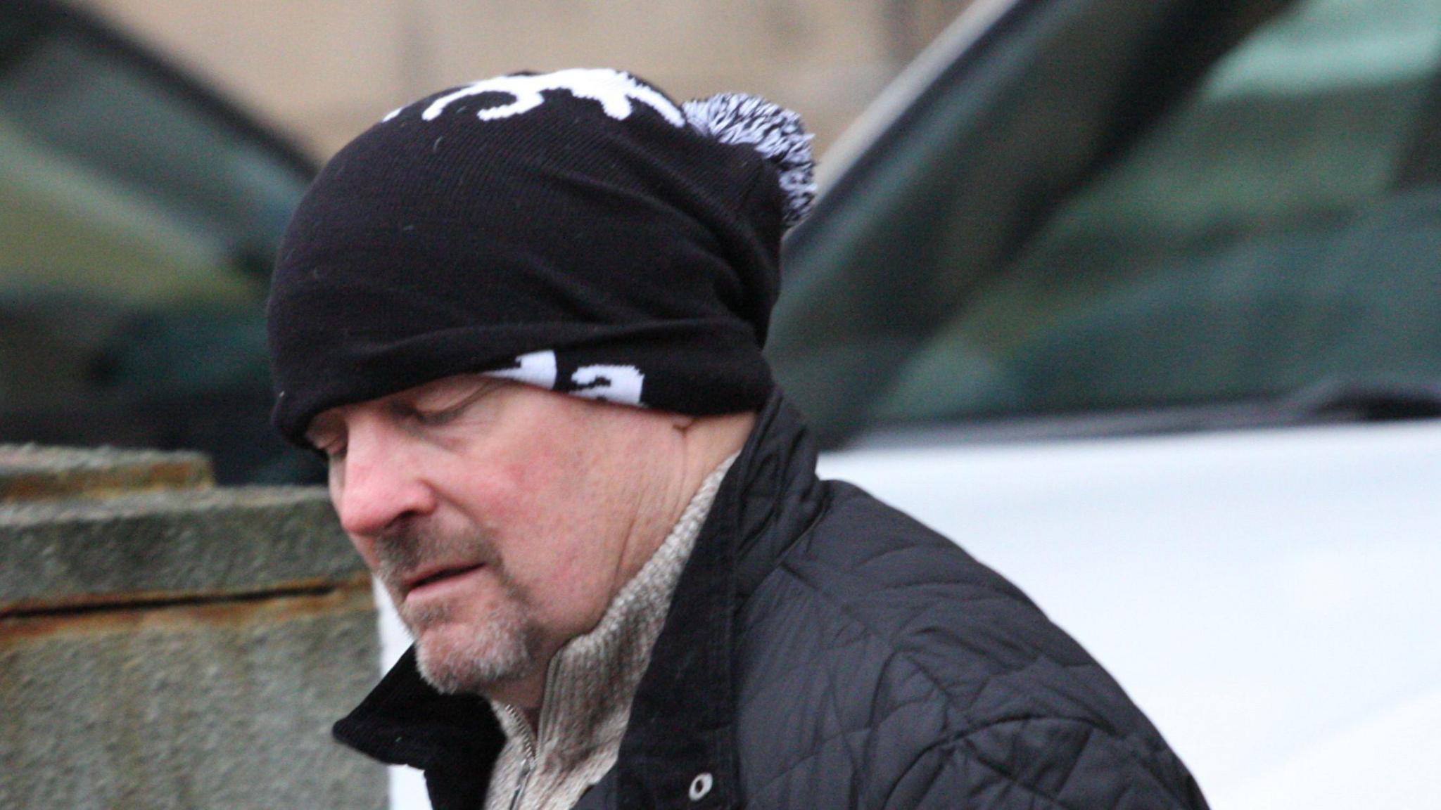 Kevin Dolan at court, wearing a black jacket and a black hat covering part of his head. He has a moustache and trim beard.