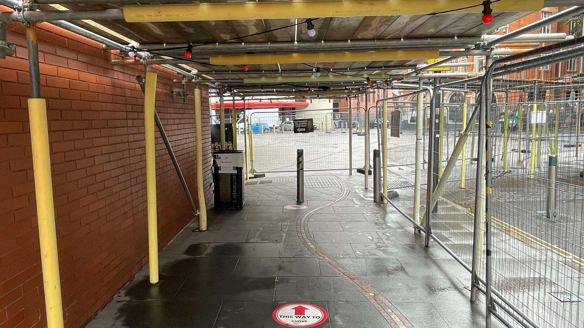 A narrow passage between a red-brick wall and safety fencing with scaffolding as a roof and yellow foam covers around the scaffolding poles