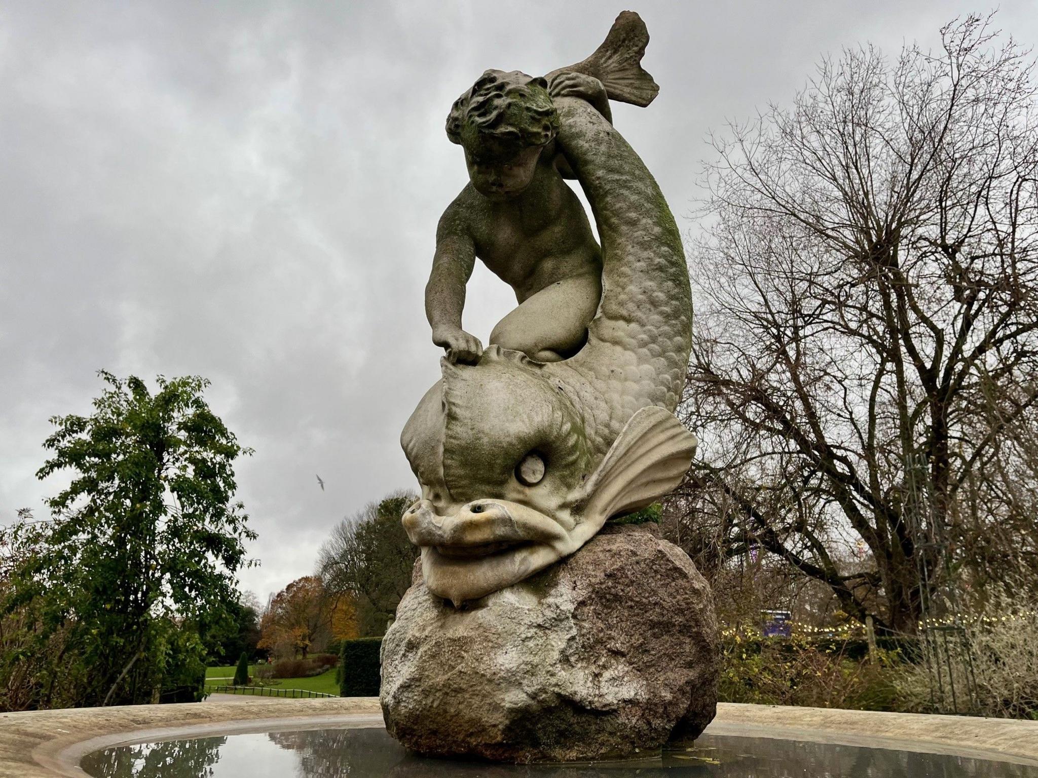 Stone sculpture of a child kneeling on the head of a sea creature on a boulder with trees and grass in the background