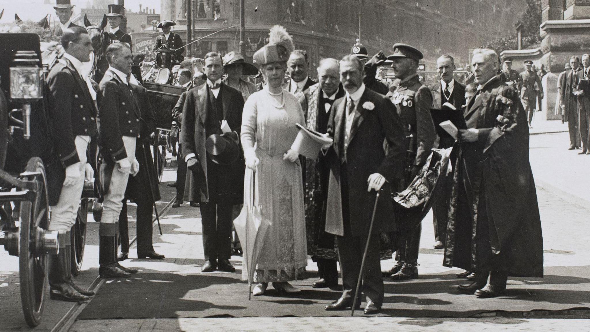 A black and white photo of King George the Fifth and Queen Mary standing outside the Wills Memorial Building having just got out of a horse and carriage, they wear formal dress and the king carries a cane. They are surrounded by dignitaries and soldiers.