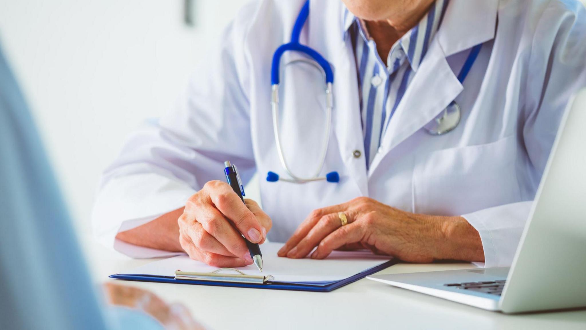 Senior female doctor writing on clipboard during discussion with male patient. Doctor and man sitting at the desk. Close up of hands, unrecognizable people.